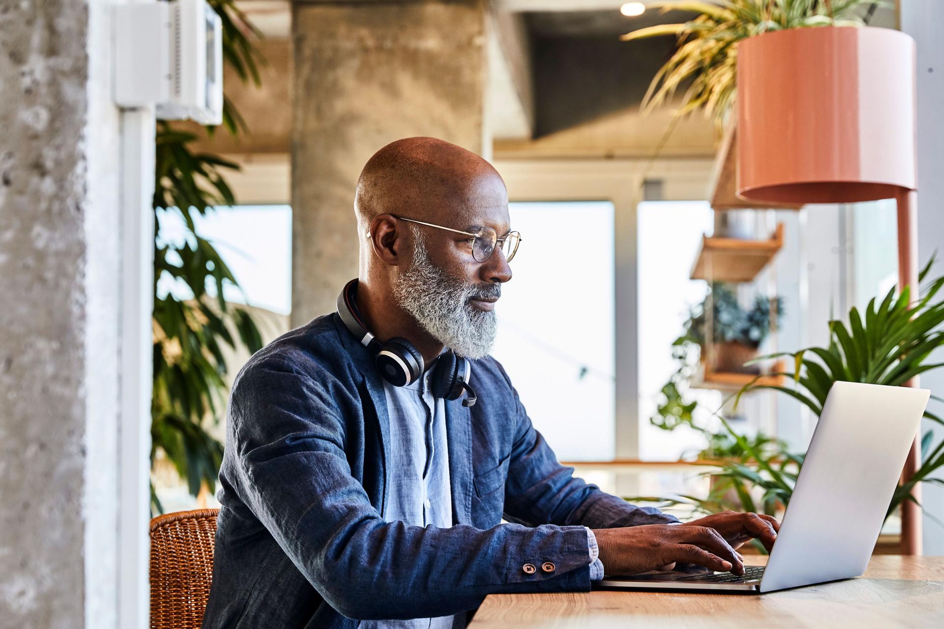 Mann mittleren Alters, der an einem Holztisch sitzt und an einem Laptop arbeitet. Im Hintergrund sind viele Pflanzen.