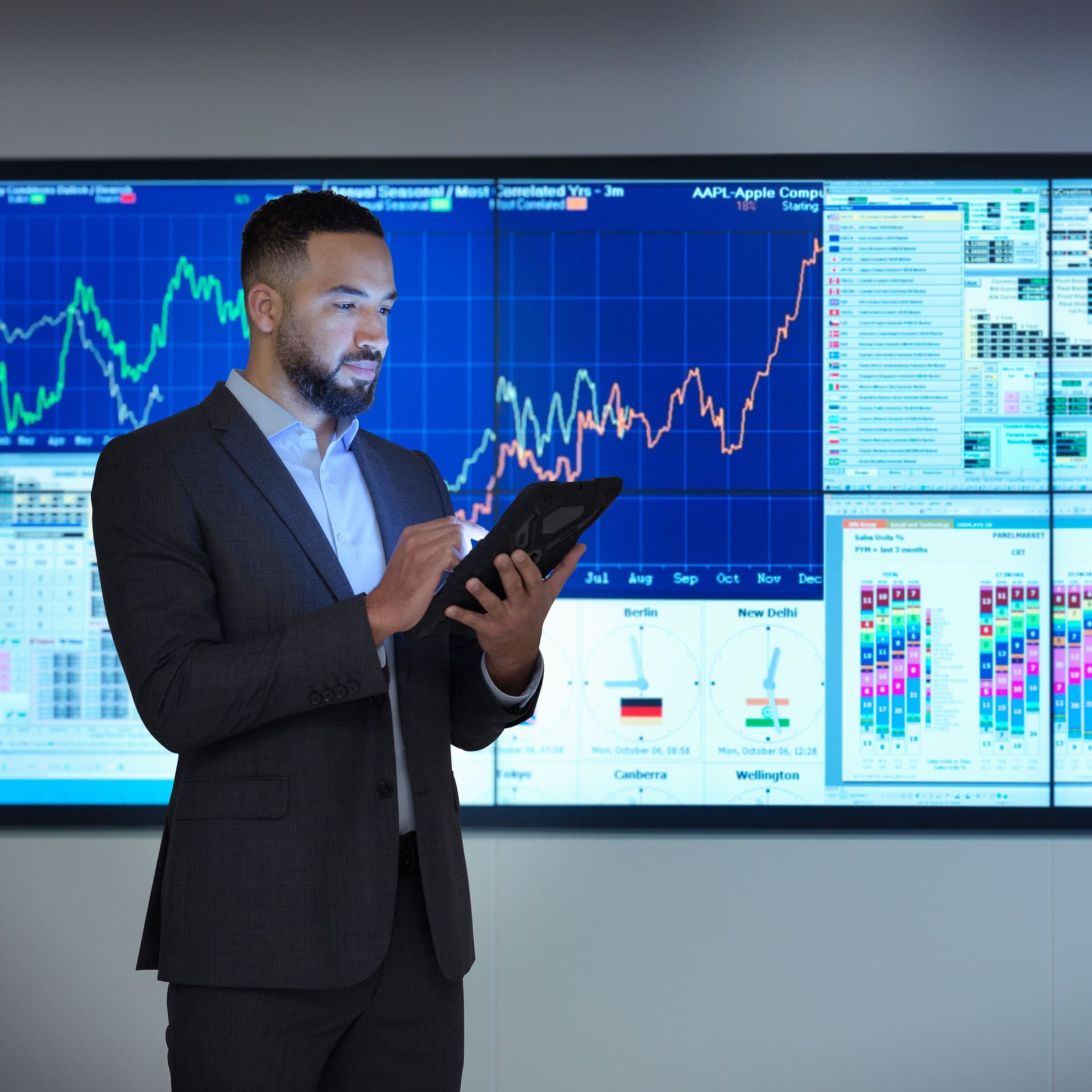 Ein Mann schaut auf sein Tablet. Im Hintergrund sind Bildschirme mit Börsenkursen zu sehen.