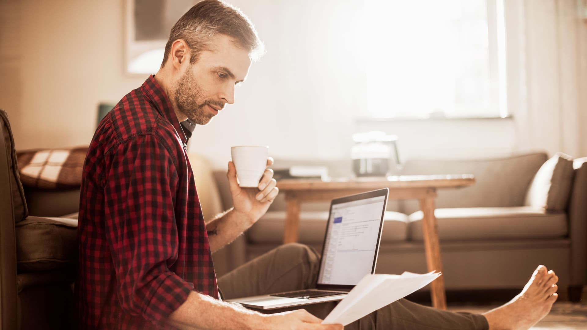 Mann sitzt auf dem Sofa mit Laptop auf dem Schoß und sieht sich unterlagen an