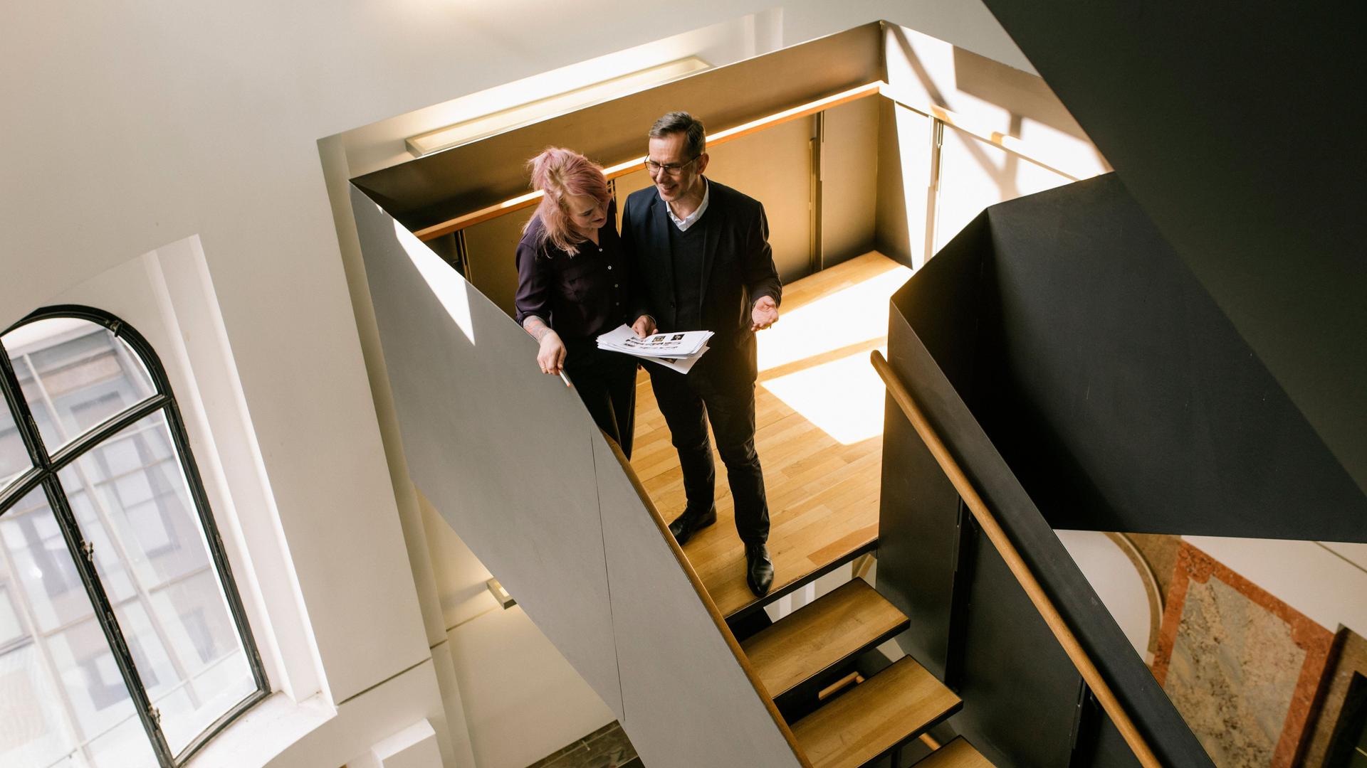 Ein Mann und eine Frau unterhalten sich in einem modernen Treppenhaus auf einer Holztreppe und gucken dabei auf Dokumente.