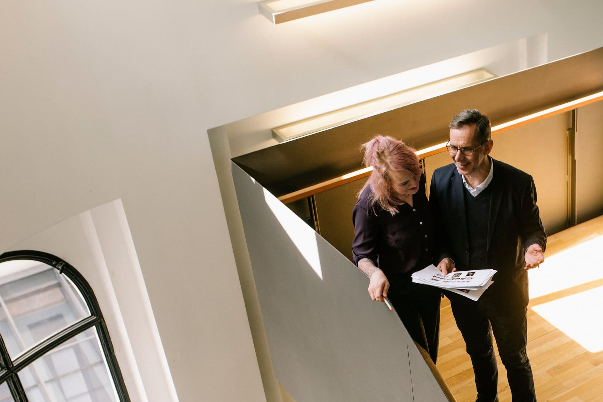 Ein Mann und eine Frau unterhalten sich in einem modernen Treppenhaus auf einer Holztreppe und gucken dabei auf Dokumente.