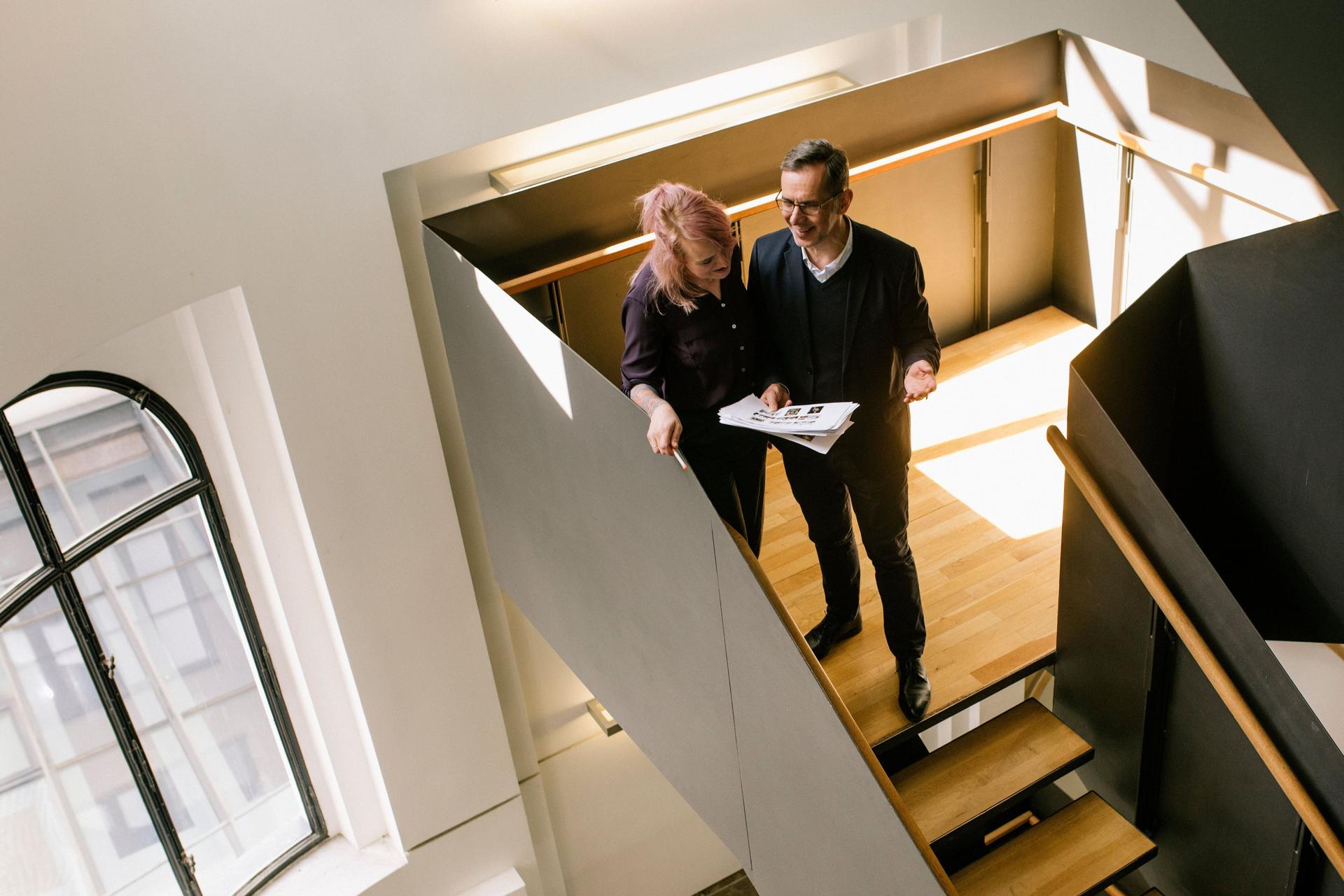 Ein Mann und eine Frau unterhalten sich in einem modernen Treppenhaus auf einer Holztreppe und gucken dabei auf Dokumente.
