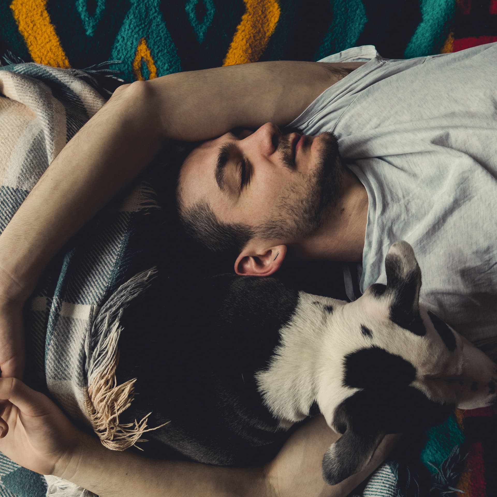 Junger Mann liegt mit Händen um einen Hund gelegt auf dem Bett und hat die Augen geschlossen. Blick von Oben.