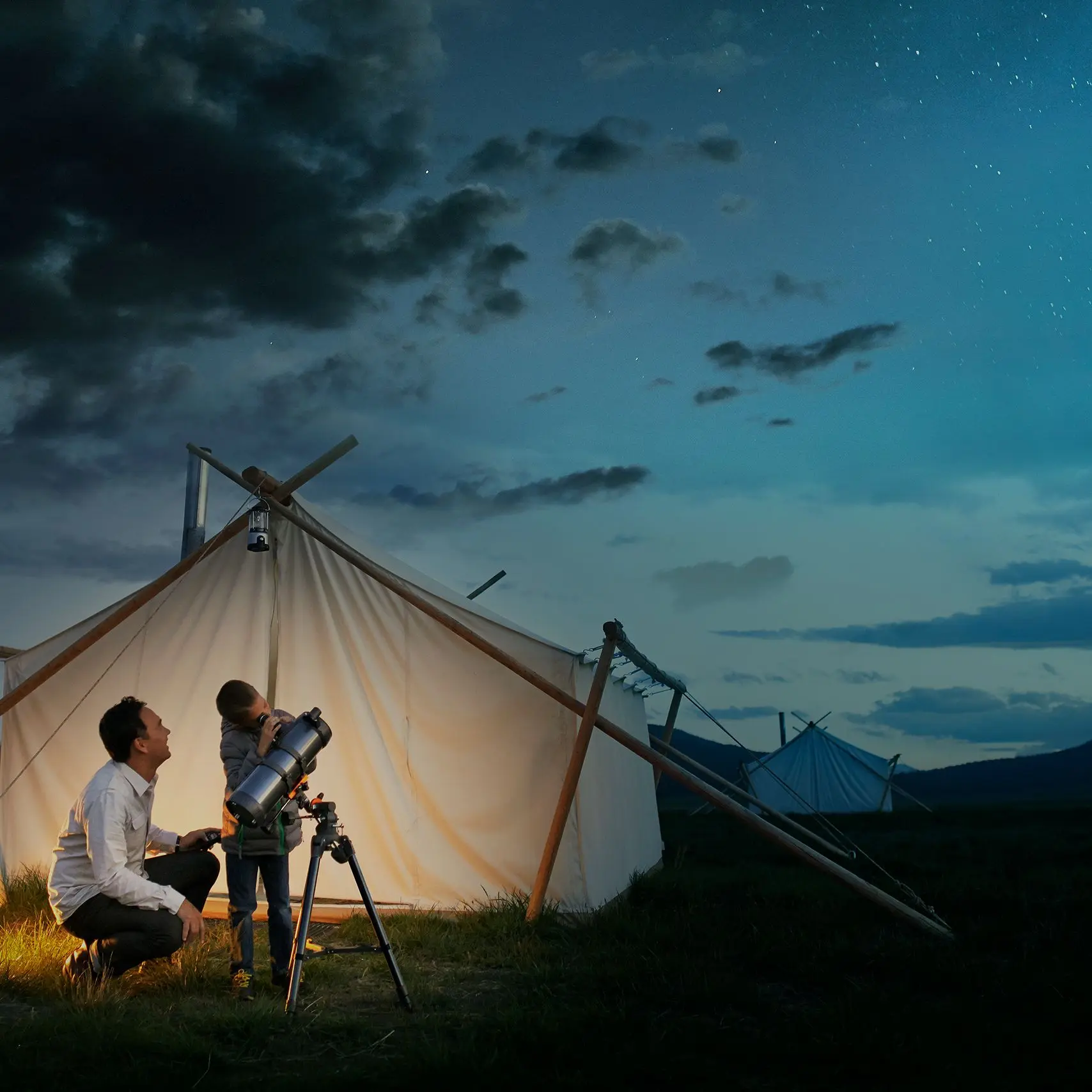 Ein Mann und ein Kind stehen abends vor einem beleuchteten Zelt auf einer Wiese und betrachten gemeinsam den Sternenhimmel durch ein Teleskop. Die Szene wirkt ruhig und idyllisch, umgeben von weiteren Zelten und einem leicht bewölkten Nachthimmel.