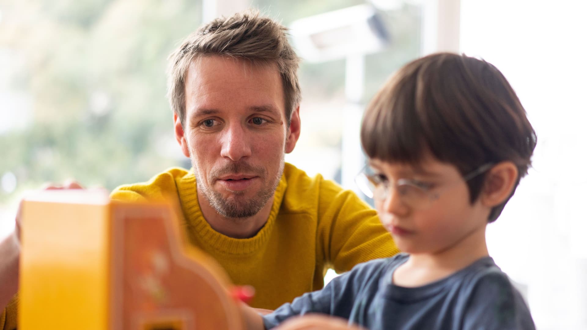 Ein Mann mit gelbem Pullover und ein Kind mit Brille spielen mit einer Spielzeugkasse.