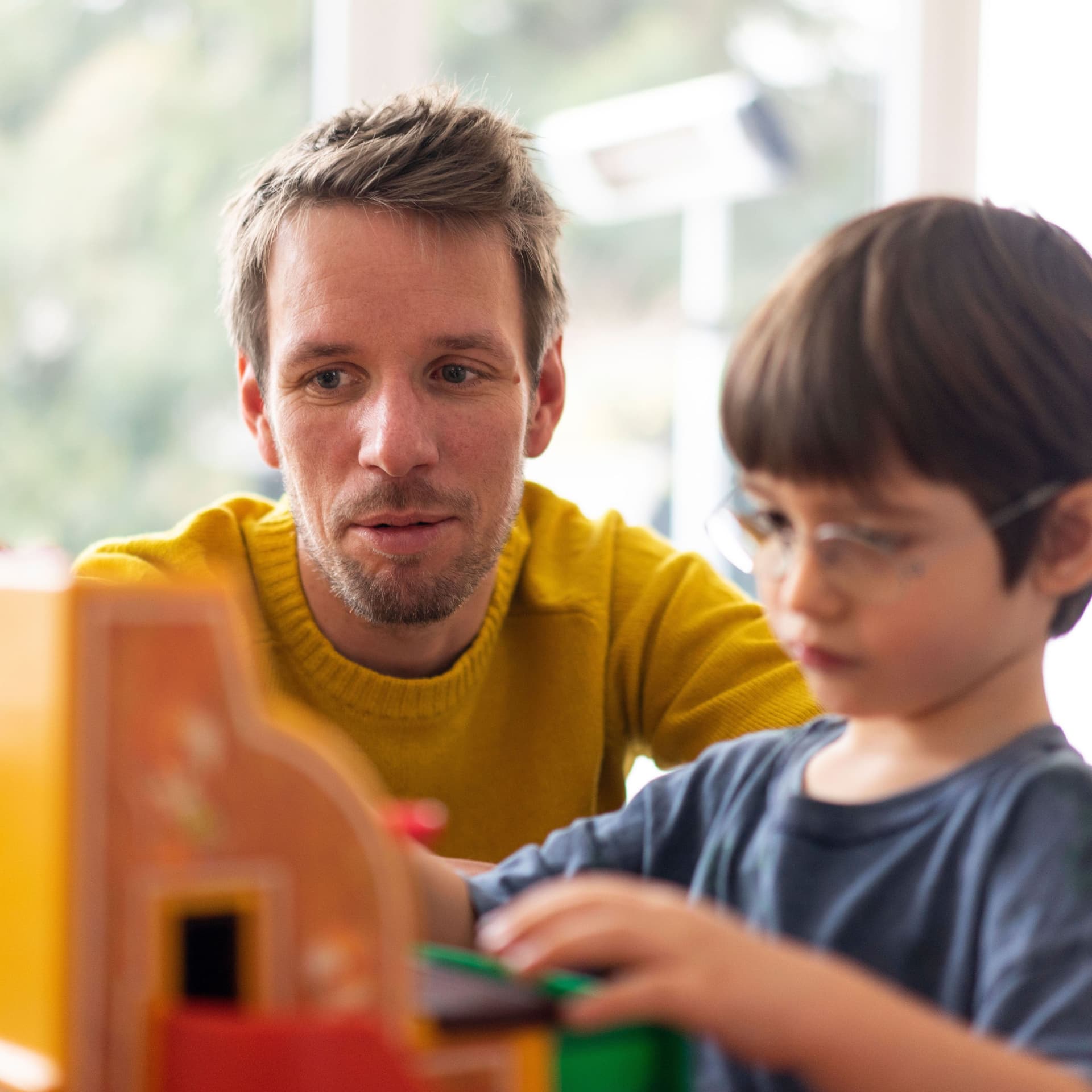 Ein Mann mit gelbem Pullover und ein Kind mit Brille spielen mit einer Spielzeugkasse.