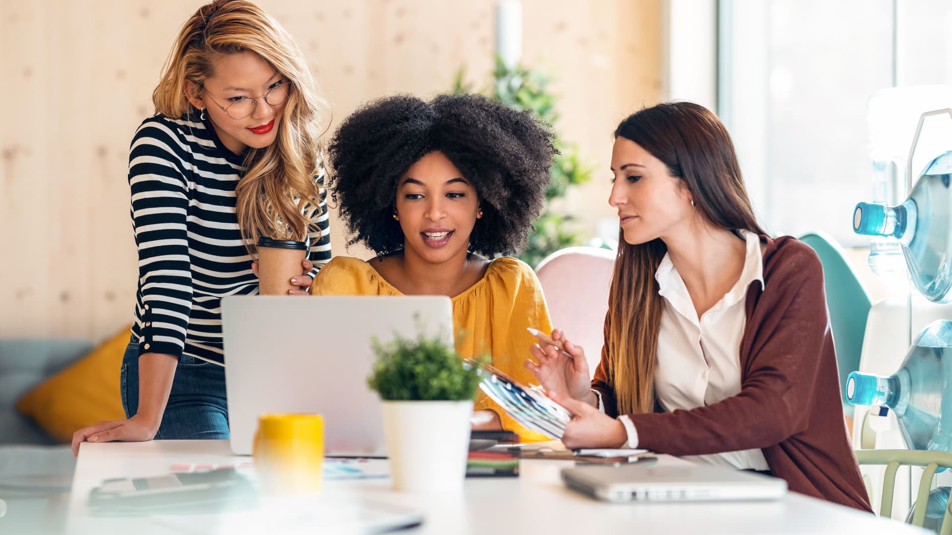 Drei junge Frauen arbeiten zusammen an einem Laptop. Sie sitzen und stehen an einem Tisch in einem bunt eingerichteten Büro.