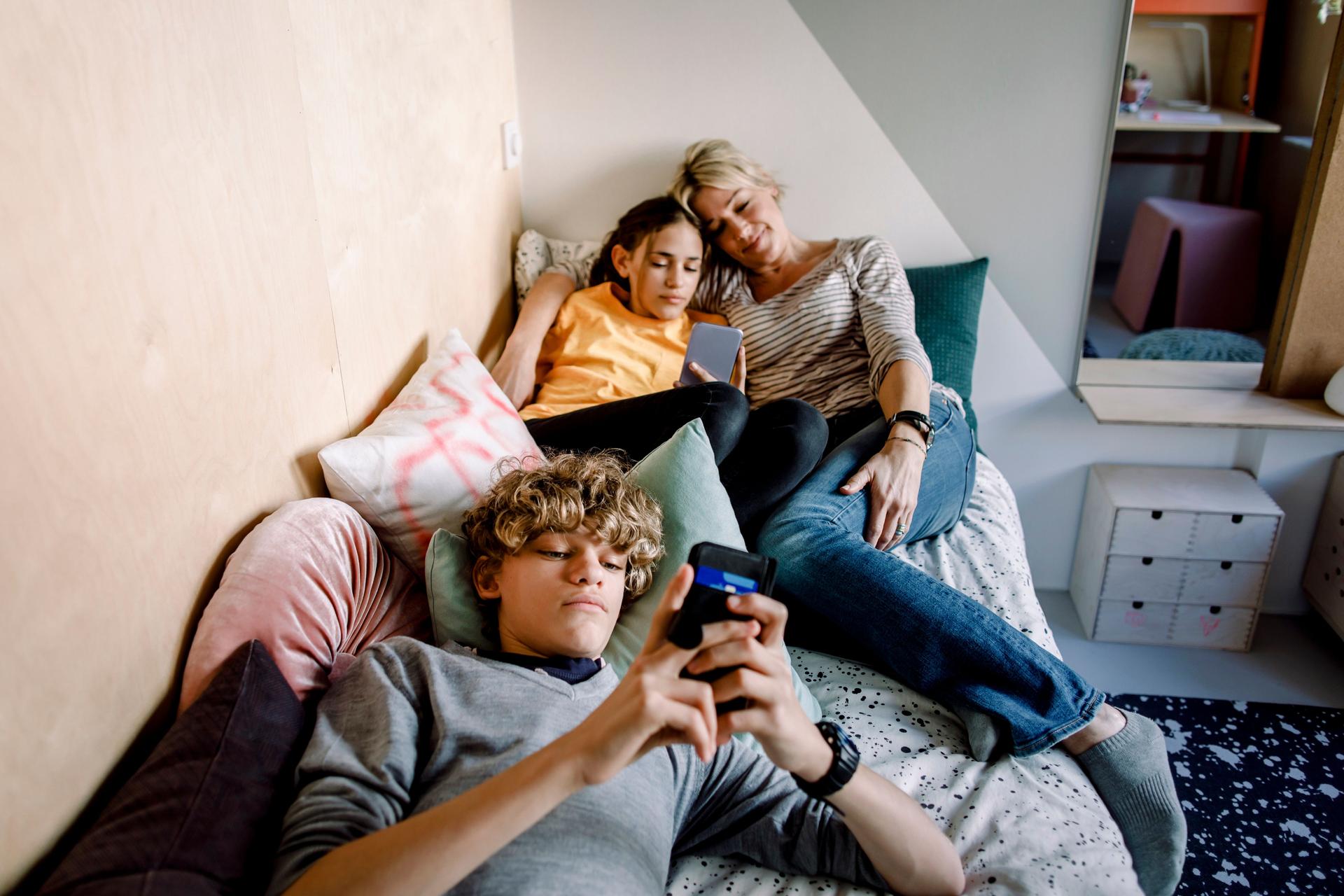 Mutter liegt mit Tochter und Sohn zusammen in einem Bett während diese am Handy sind.