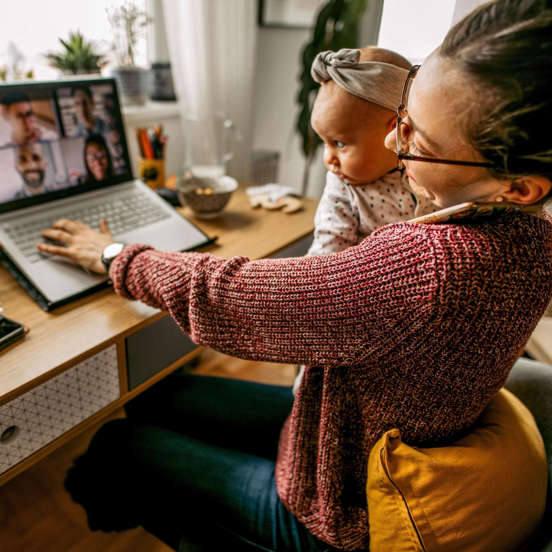 Junge Mutter mit Baby auf dem Schoß sitzt an ihrem Schreibtisch zu Hause und nimmt an einer Videokonferenz teil.