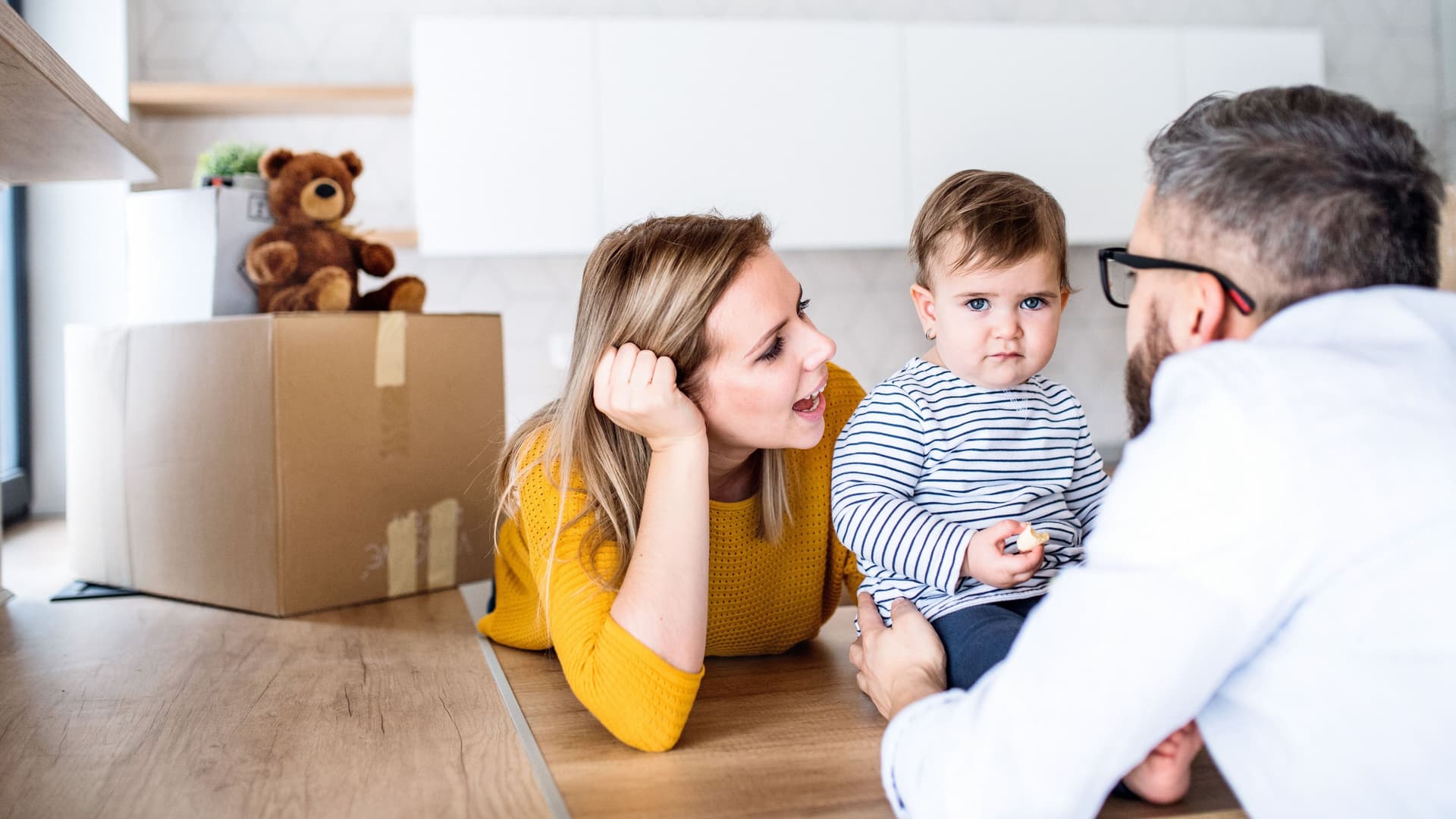 Ein Porträt einer jungen Familie mit einem Kleinkind, dass auf einem Holztisch sitzt. Es wird gehalten von dem Vater, der mit dem Rücken zur Kamera steht.