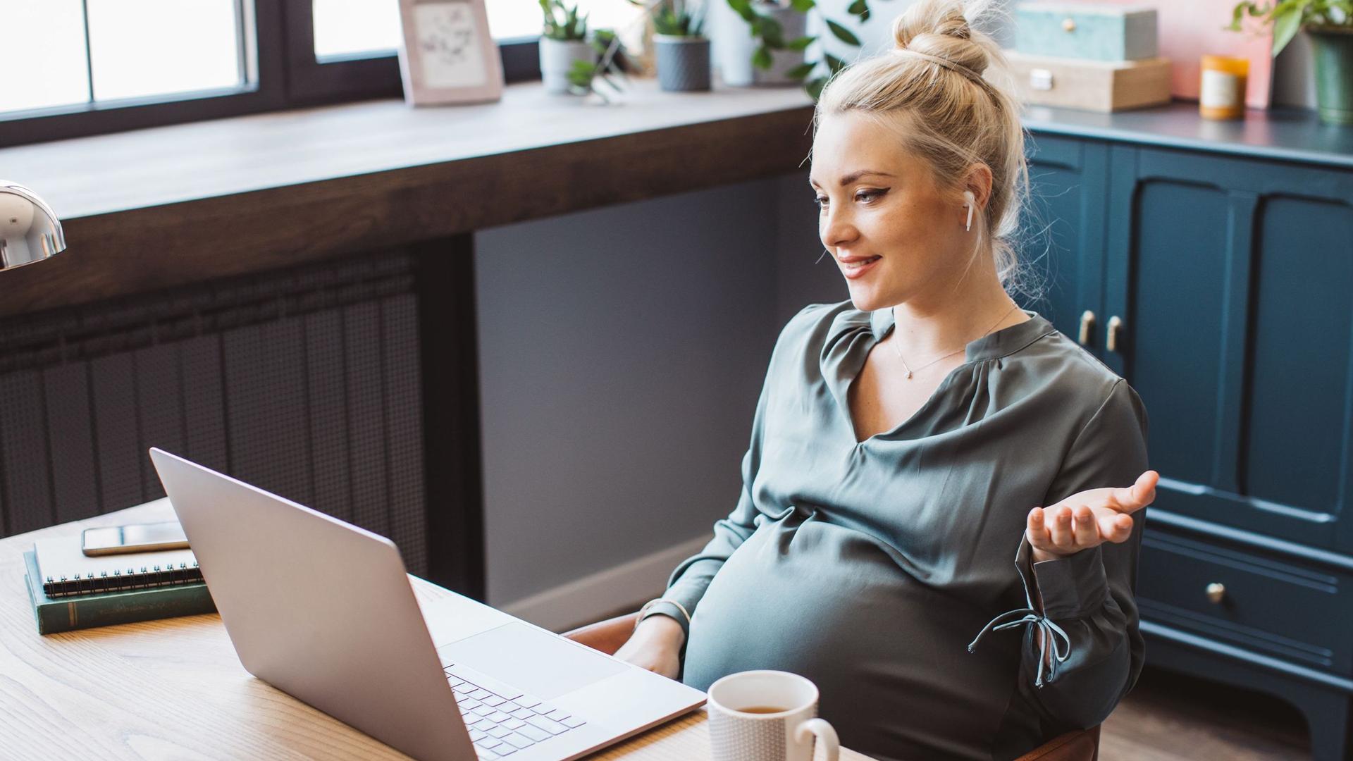 Eine hochschwangere junge Frau in Business-Kleidung sitzt entspannt gestikulierend mit Ear-In-Kopfhörern vor einem Laptop und hält einen Videocall. 