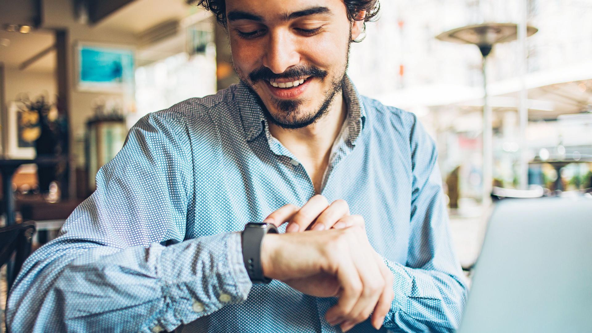 Portrait eines jungen Mannes, der in einem Café auf seine Smartwatch blickt.