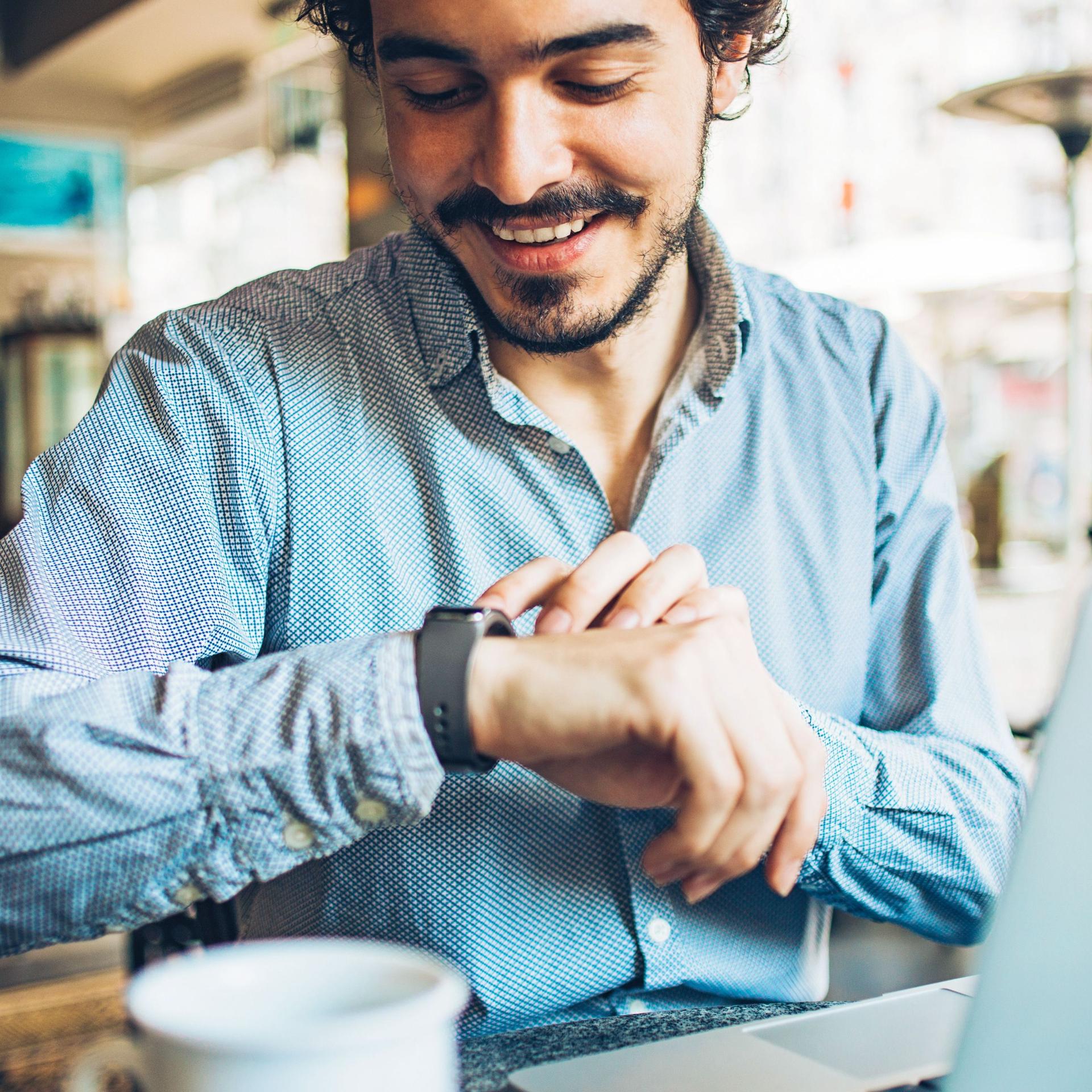 Portrait eines jungen Mannes, der in einem Café auf seine Smartwatch blickt.