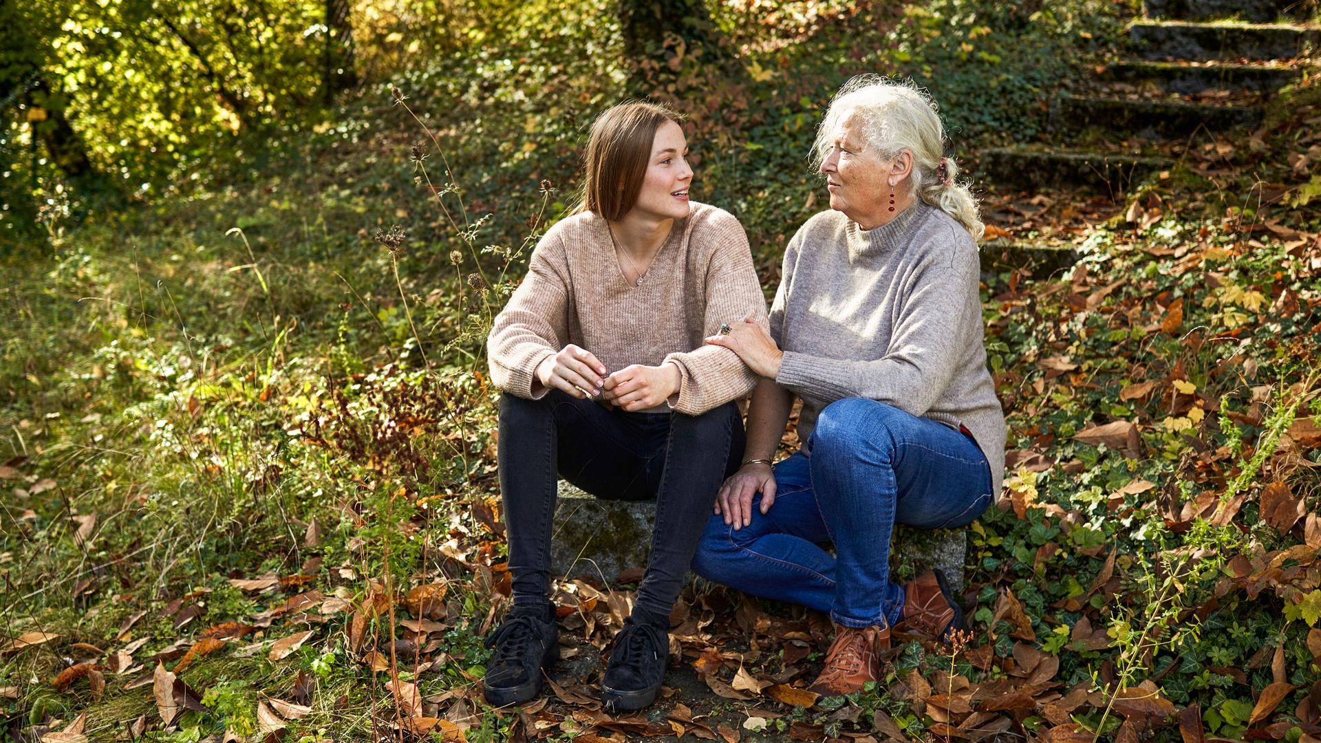 Eine ältere Frau und eine jüngere Frau sitzen auf einer Treppe , die von Herbstlaub bedeckt ist.