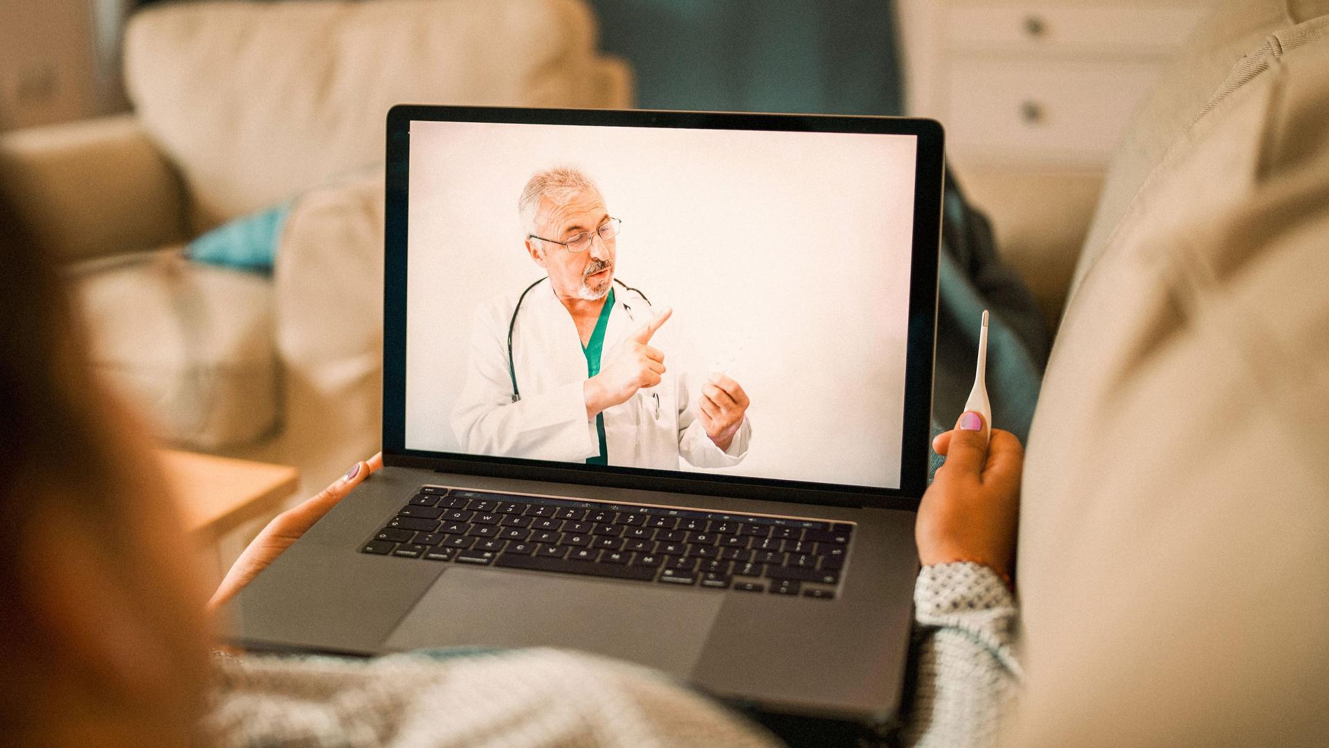 Eine kranke Frau führt ein Videogespräch mit einem Arzt. Sie hält ein Fieberthermometer in der Hand.