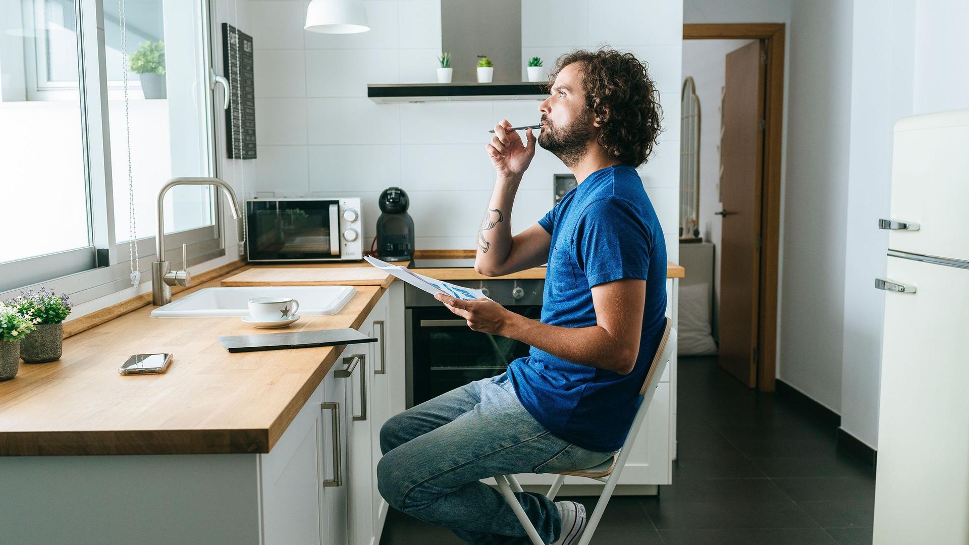 Ein Mann in einem blauen T-SHirt sitzt in seiner Küche mit mehreren Papieren in der Hand und denkt nach.