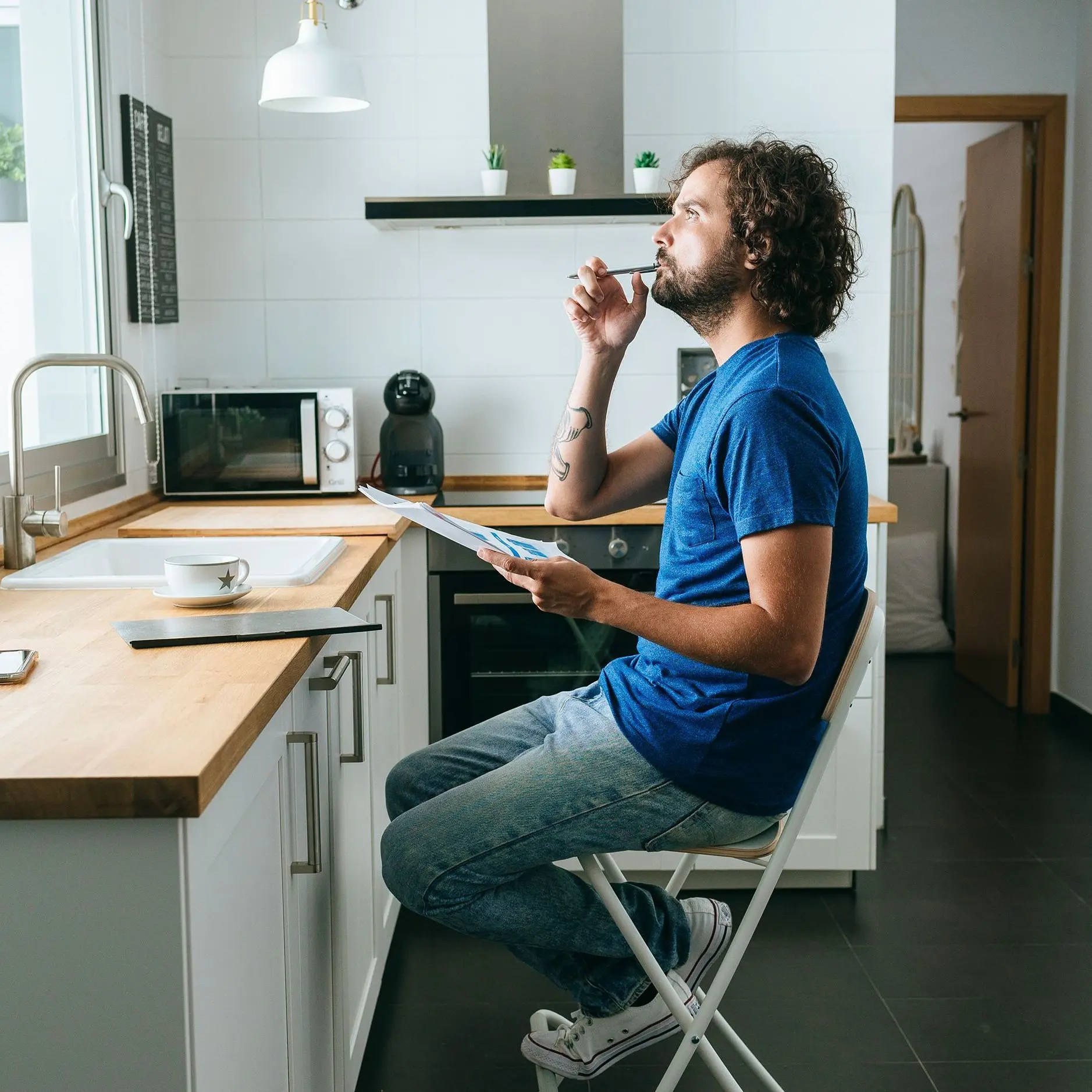 Ein Mann in einem blauen T-SHirt sitzt in seiner Küche mit mehreren Papieren in der Hand und denkt nach.