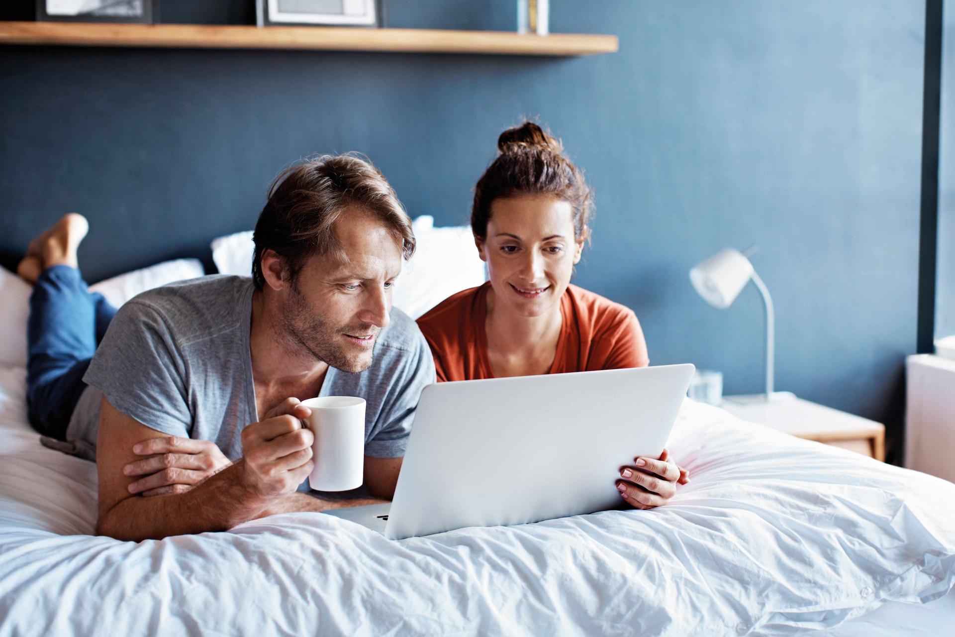 Ein Mann und eine Frau mittleren Alters liegen bäuchlings auf einem Bett. Sie schauen gemeinsam auf einen Laptopbildschirm.