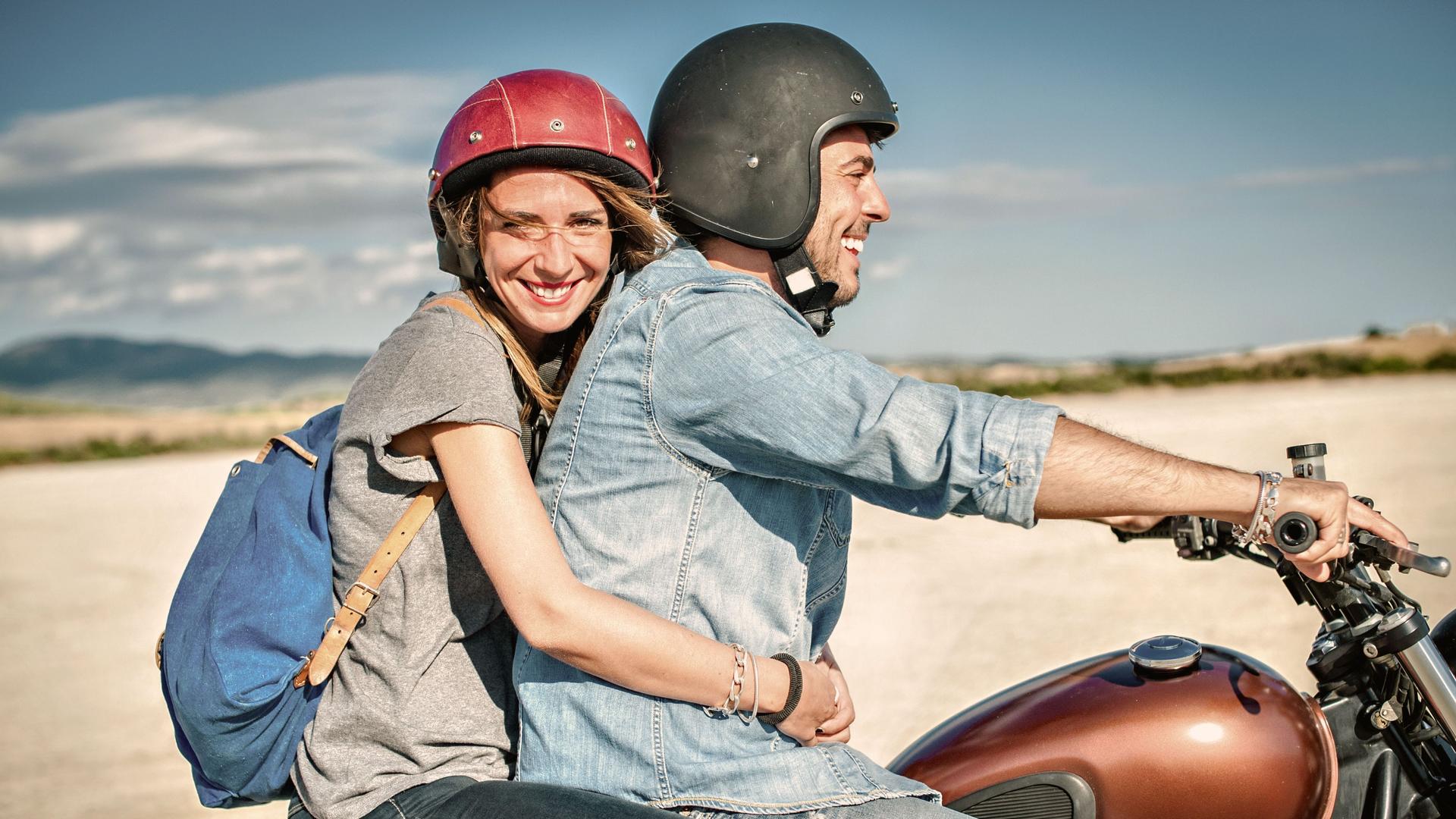 Frau und Mann fahren auf einem Motorrad am Strand. Sie lächelt in die Kamera.