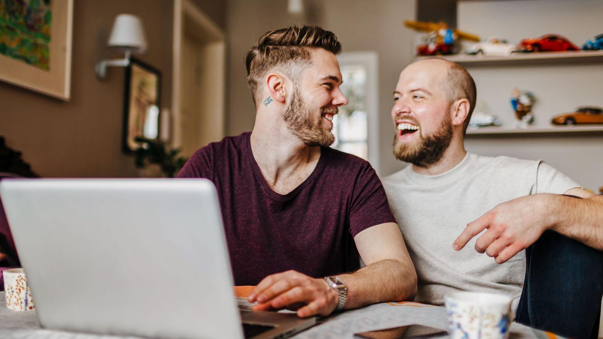 Zwei junge Männer sitzen vor einem Laptop in einer Wohnung. Sie schauen sich an und lachen ausgelassen.