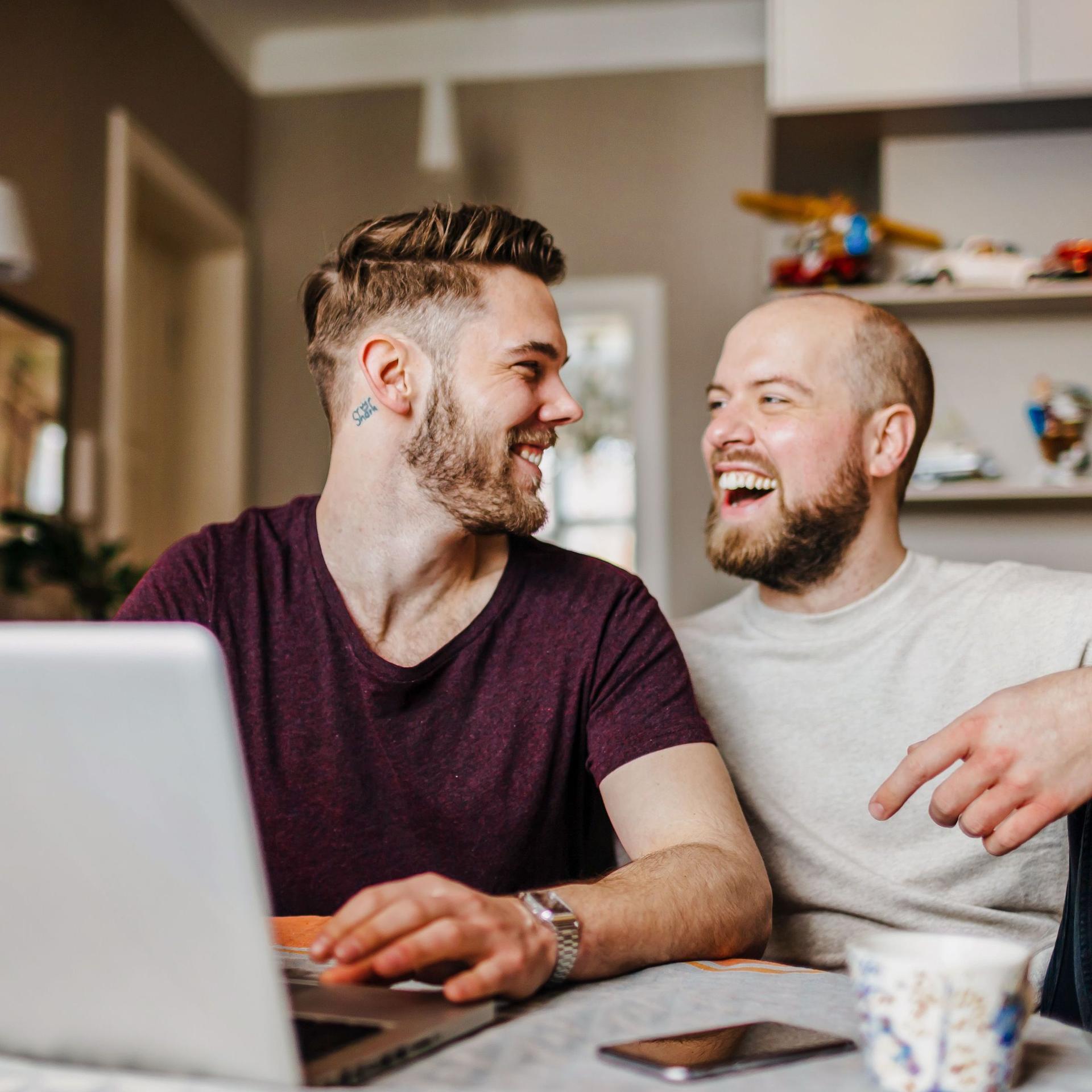 Zwei junge Männer sitzen vor einem Laptop in einer Wohnung. Sie schauen sich an und lachen ausgelassen.