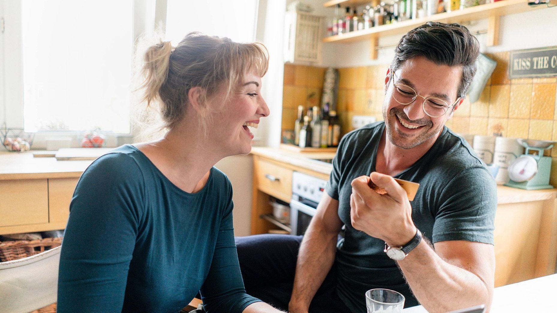 Eine Frau und ein Mann sitzen in einer gemütlichen Küche und Lachen. Beide schauen auf eine Bankkarte und lachen ausgelassen.