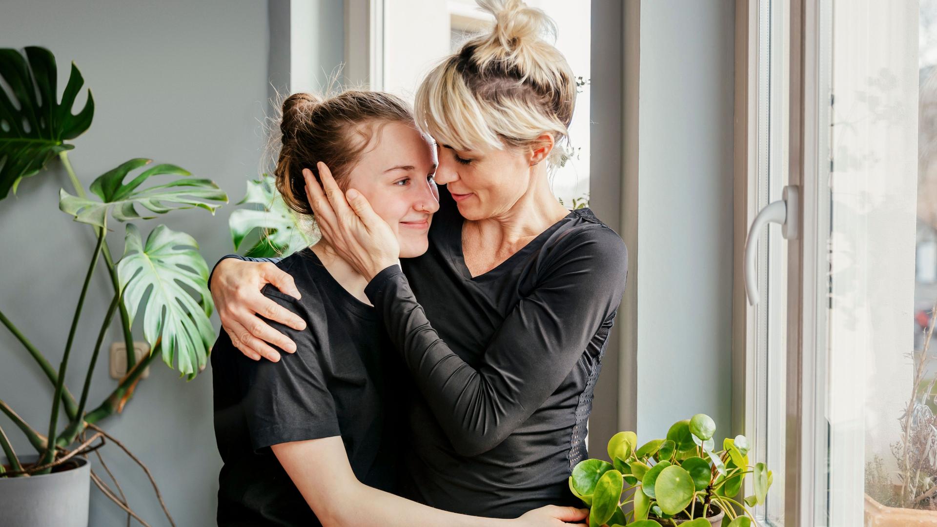 Eine Mutter steht mit ihrer Teenager Tochter am Fenster und umarmt sie