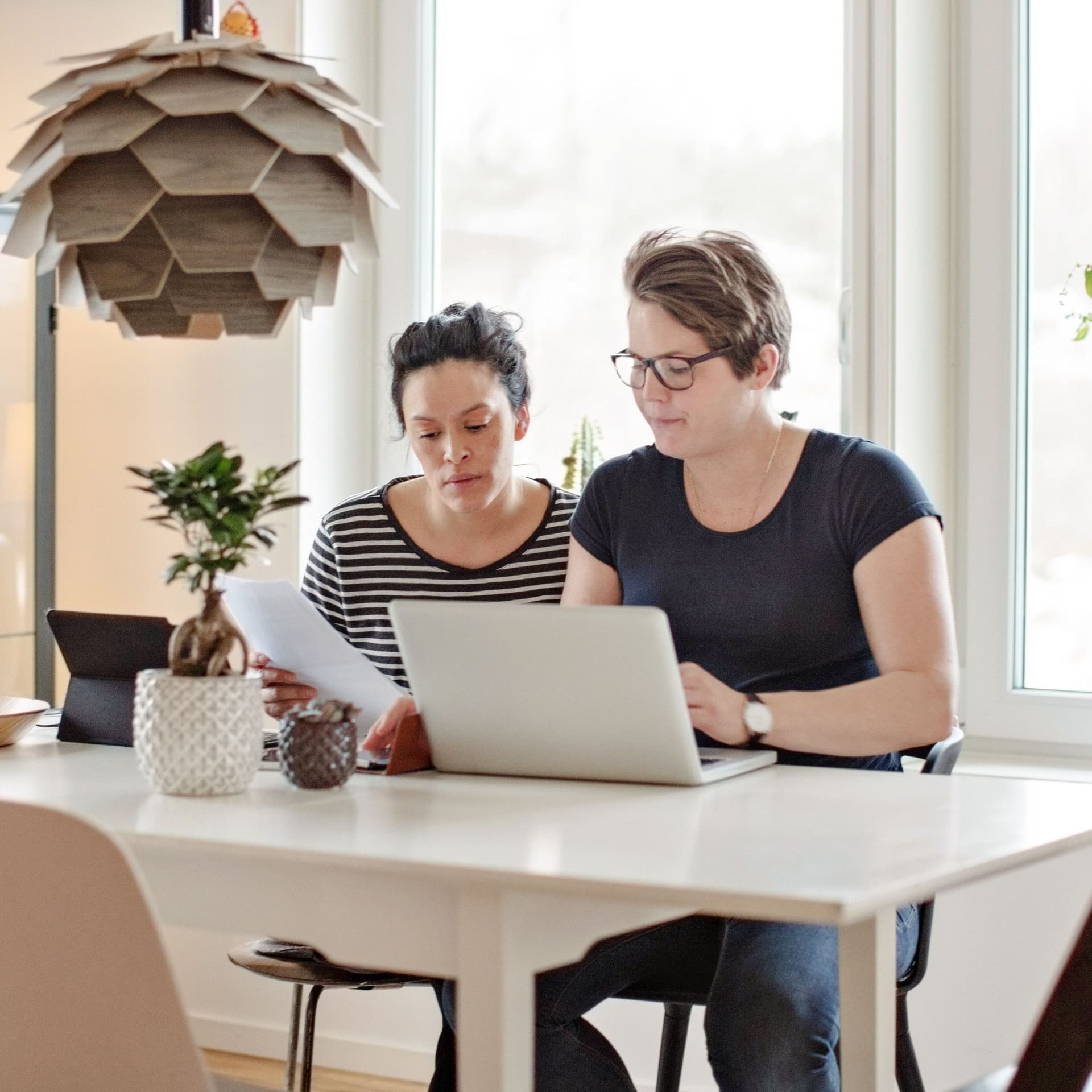 Ein gleichgeschlechtliches Paar sitzt am Wohnzimmertisch und schauen auf Dokumente. Vor den beiden Frauen steht ein Laptop.