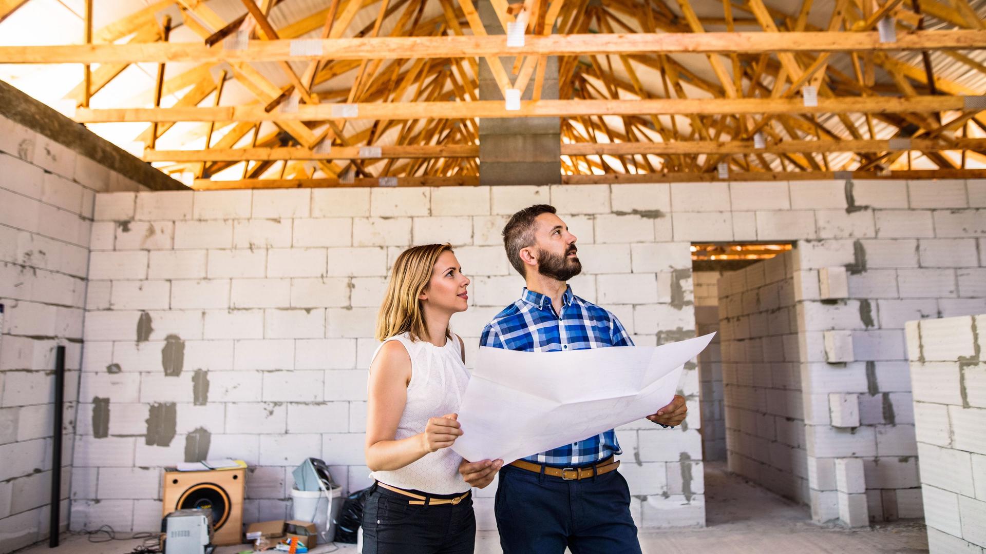 Eine Frau und ein Mann stehen in einem Neubau und halten die Pläne des Hauses in den Händen. Hinter ihnen sind neue Mauern und der Dachstuhl aus Holz.