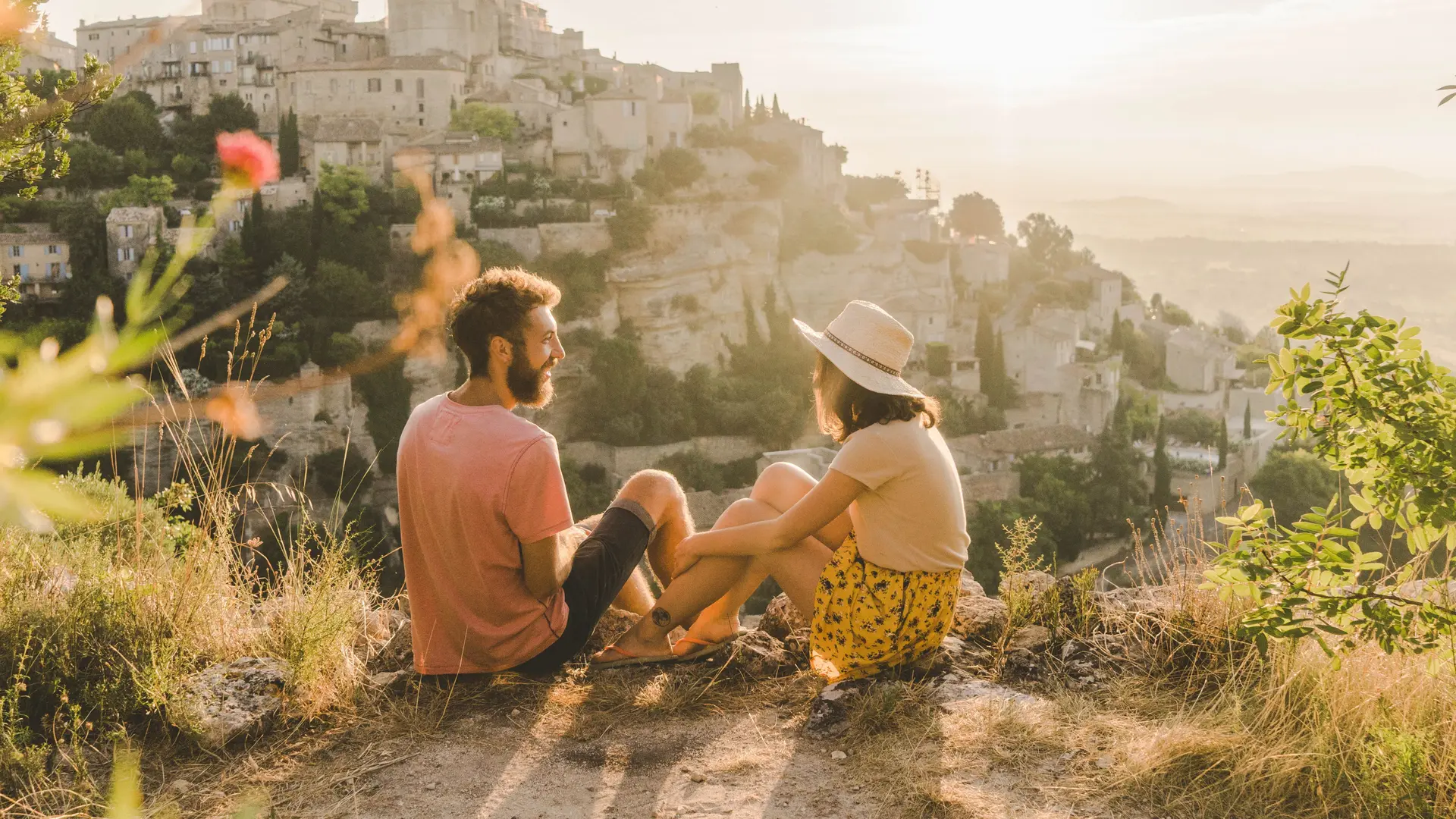 Frau und Mann sitzen an einer Klippe mit Blick auf eine malerische Stadt in der Provence. Die Sonne scheint.
