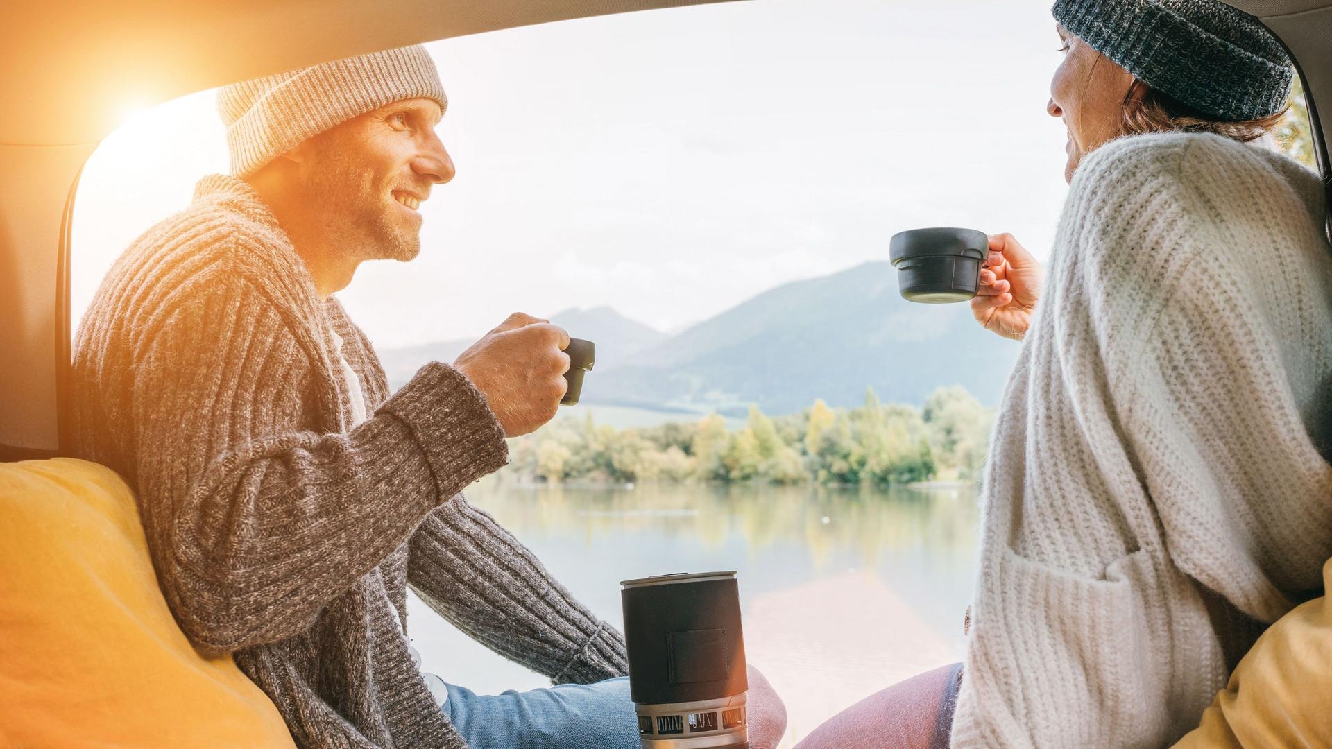 Ein Mann und eine Frau trinken Kaffee im Heck eines Campers. Beide tragen Mützen und warme Pullover.