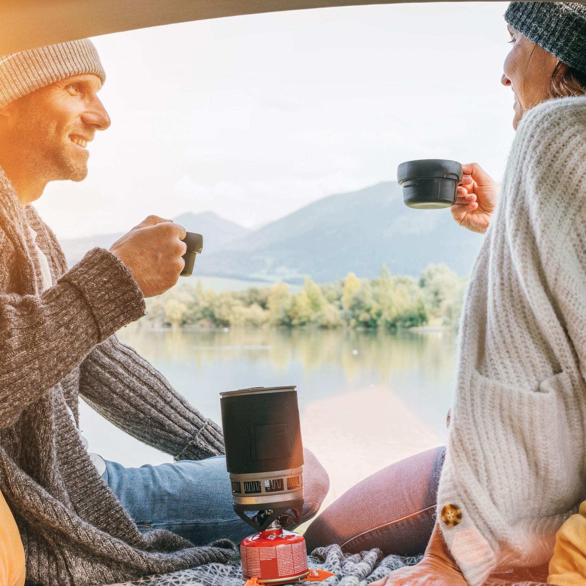 Ein Mann und eine Frau trinken Kaffee im Heck eines Campers. Beide tragen Mützen und warme Pullover.