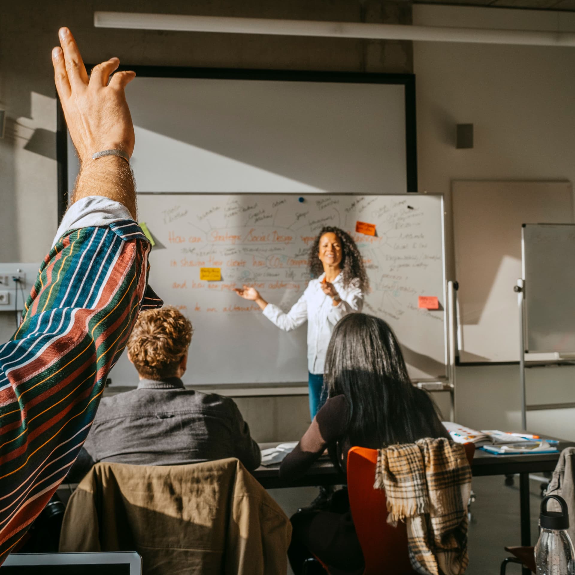 Eine lehrende Person steht an einem Whiteboard in einem Klassenraum. In ihm sitzen erwachsene Schüler, einer davon meldet sich.
