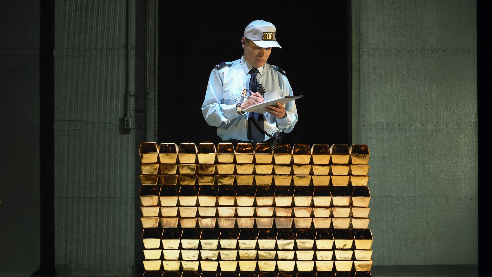 Ein Sicherheitsmitarbeiter in Uniform und Cap steht hinter einem Stapel glänzender Goldbarren und macht sich handschriftliche Notizen auf einem Klemmbrett.