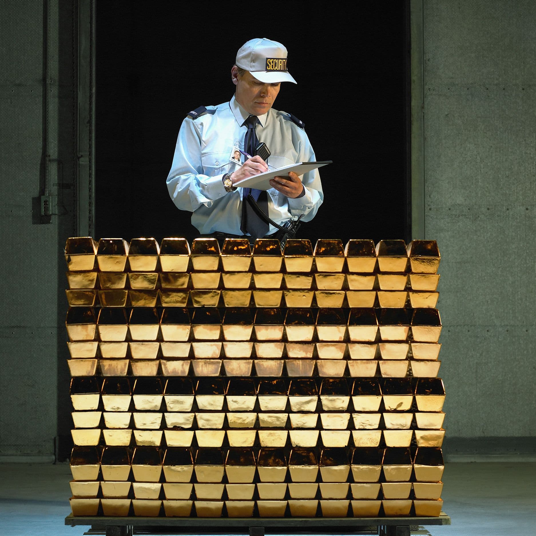 Ein Sicherheitsmitarbeiter in Uniform und Cap steht hinter einem Stapel glänzender Goldbarren und macht sich handschriftliche Notizen auf einem Klemmbrett.