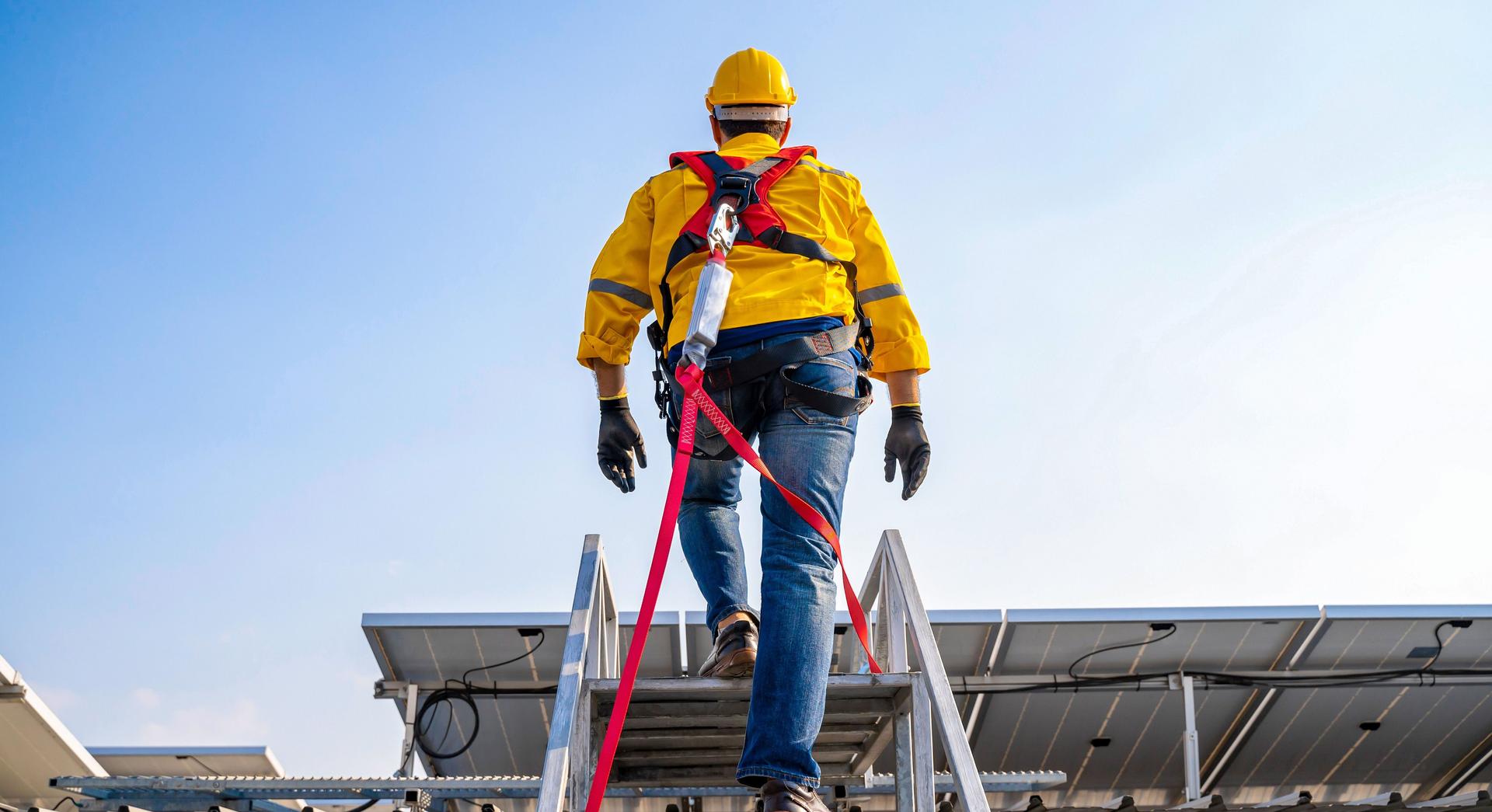 Person in gelber Arbeitsjacke und gelbem Helm steigt mit Sicherung über eine Treppe auf ein Häuserdach.