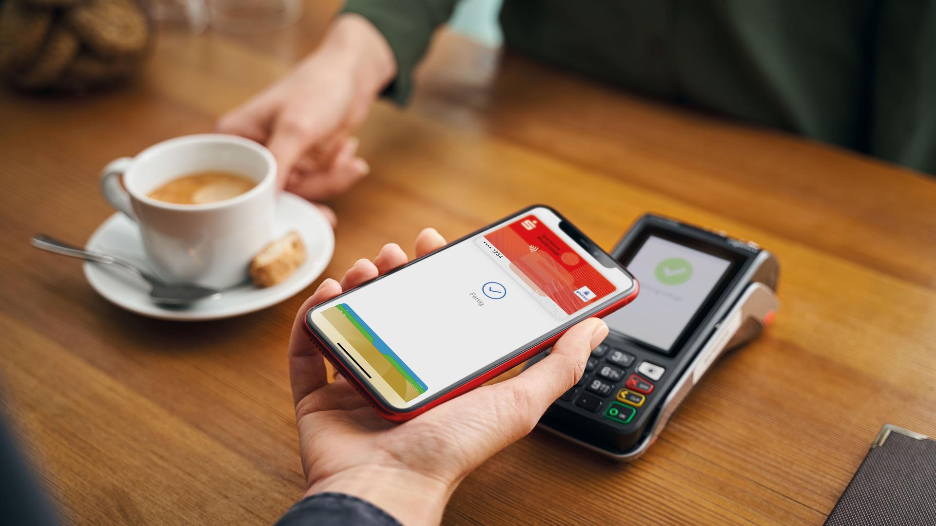 Nahaufnahe einer Hand mit Handy in einem Cafe. Auf dem Display sieht man den Bezahlvorgang für einen Kaffee über die Sparkassen-App.