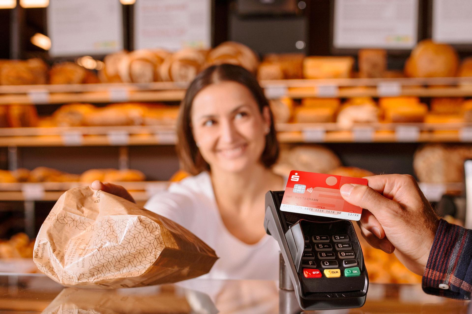 Person zahlt mit Sparkassen-Bankkarte in einer Bäckerei. Die Fachverkäuferin lächelt und reicht eine Papiertüte über den Tresen.