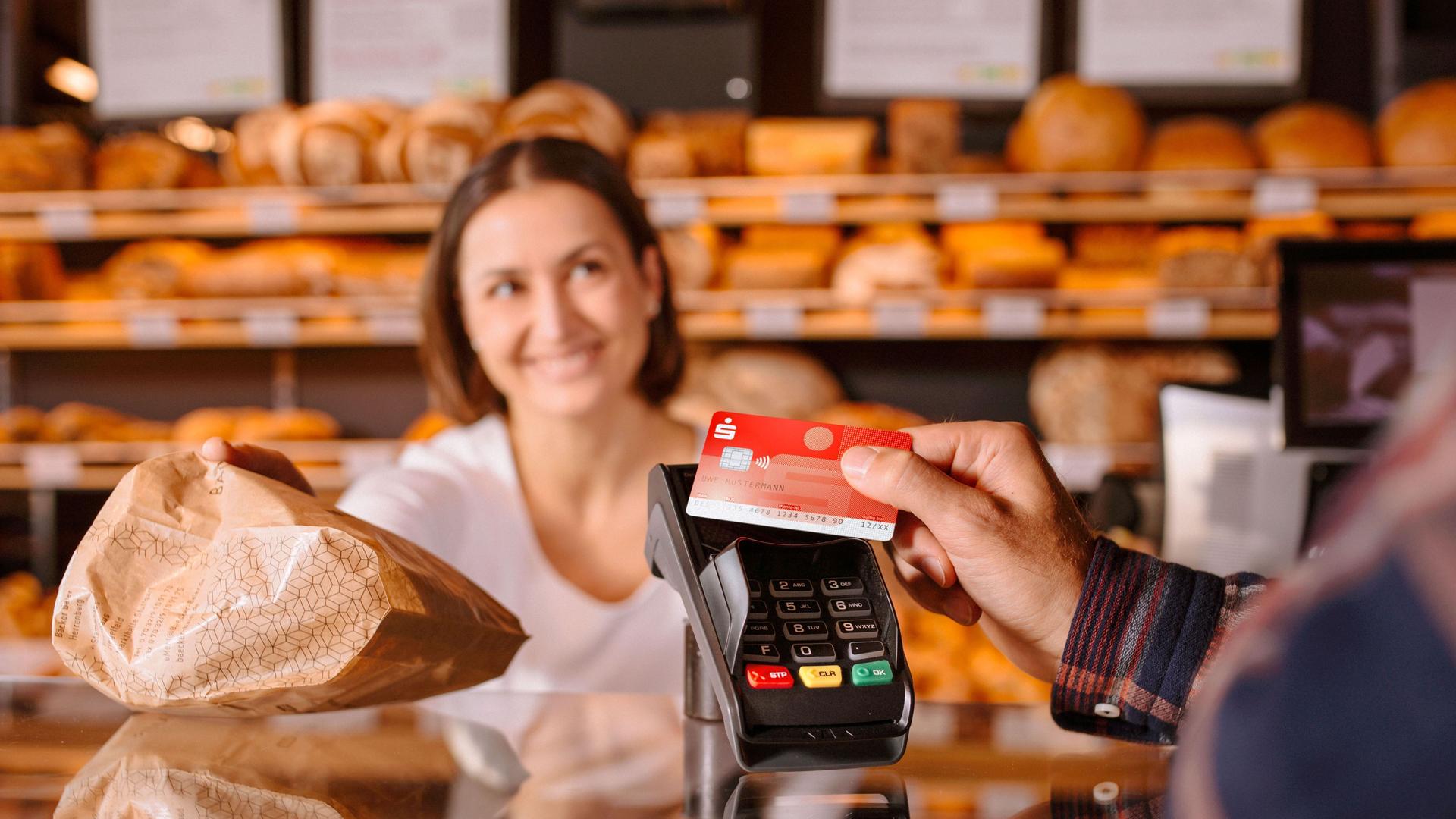Person zahlt mit Sparkassen-Bankkarte in einer Bäckerei. Die Fachverkäuferin lächelt und reicht eine Papiertüte über den Tresen.