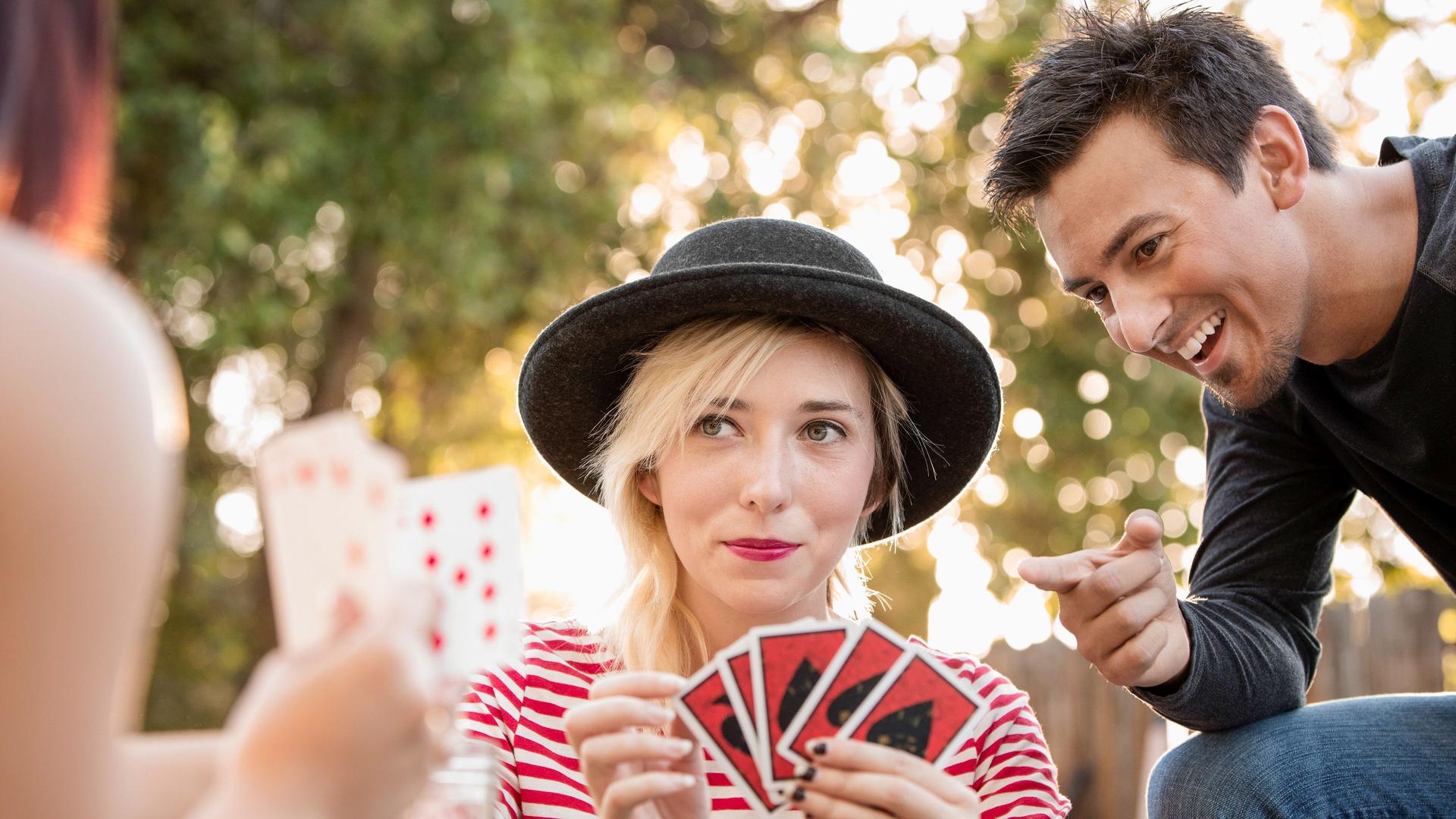Zwei junge Frauen spielen Karten im Park. Ein Mann sitzt neben ihnen und kommentiert die Karten.