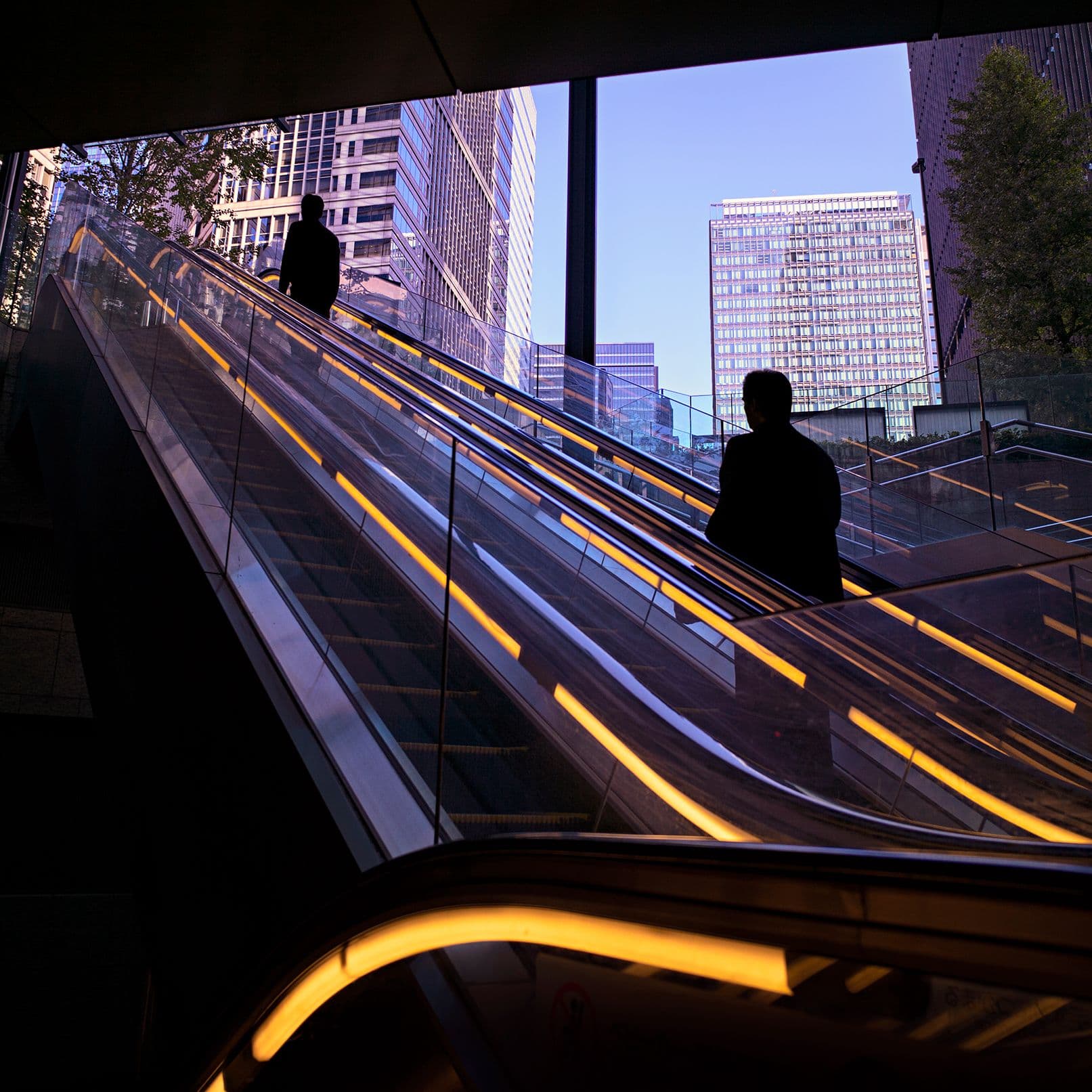Zwei Personen fahren auf Rolltreppen inmitten moderner Hochhäuser nach oben.