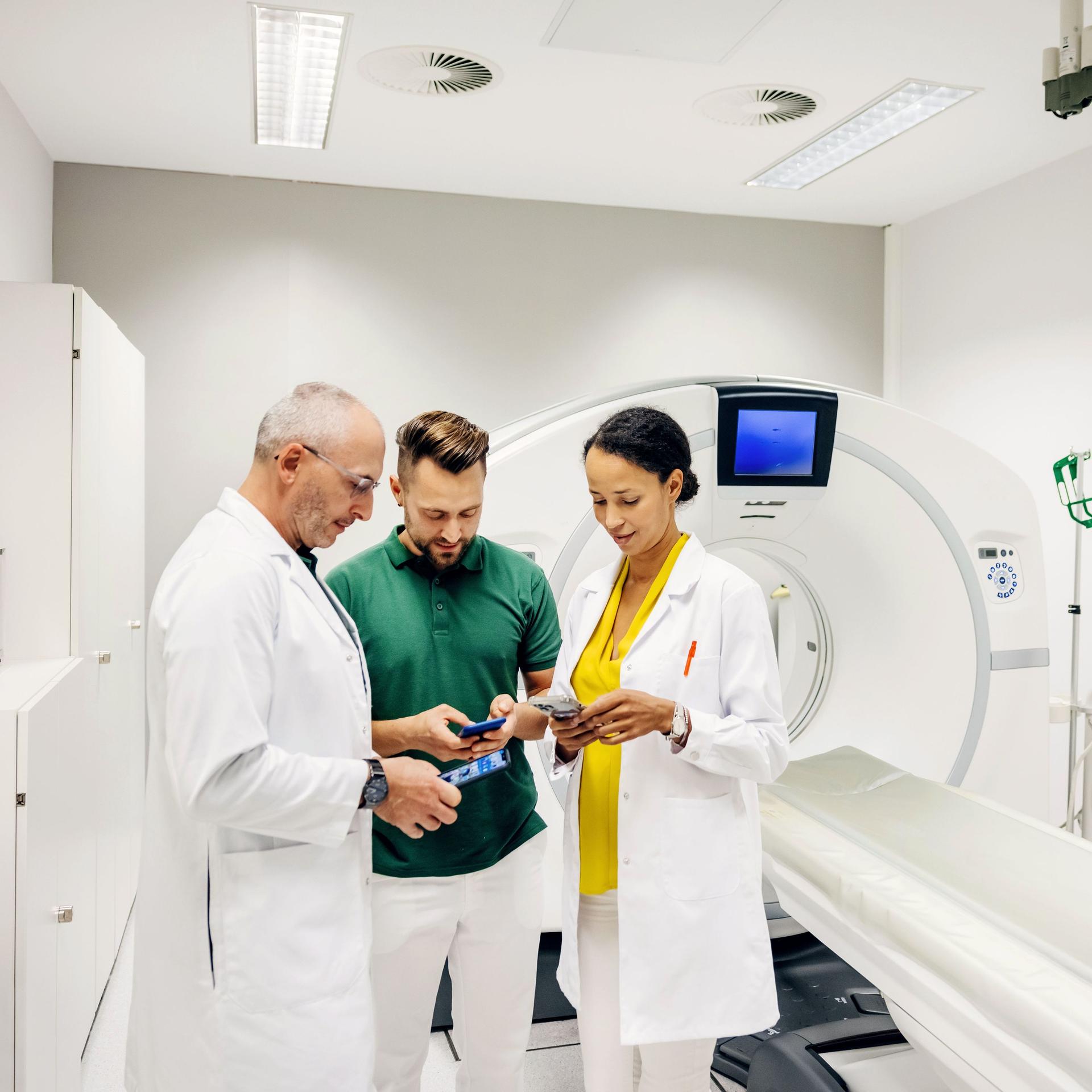 Eine Ärztin und ein Arzt besprechen einige Scandaten mit einem Mann und schauen auf ihre Smartphones, während sie in einem CT-Scanraum in einem Krankenhaus stehen.