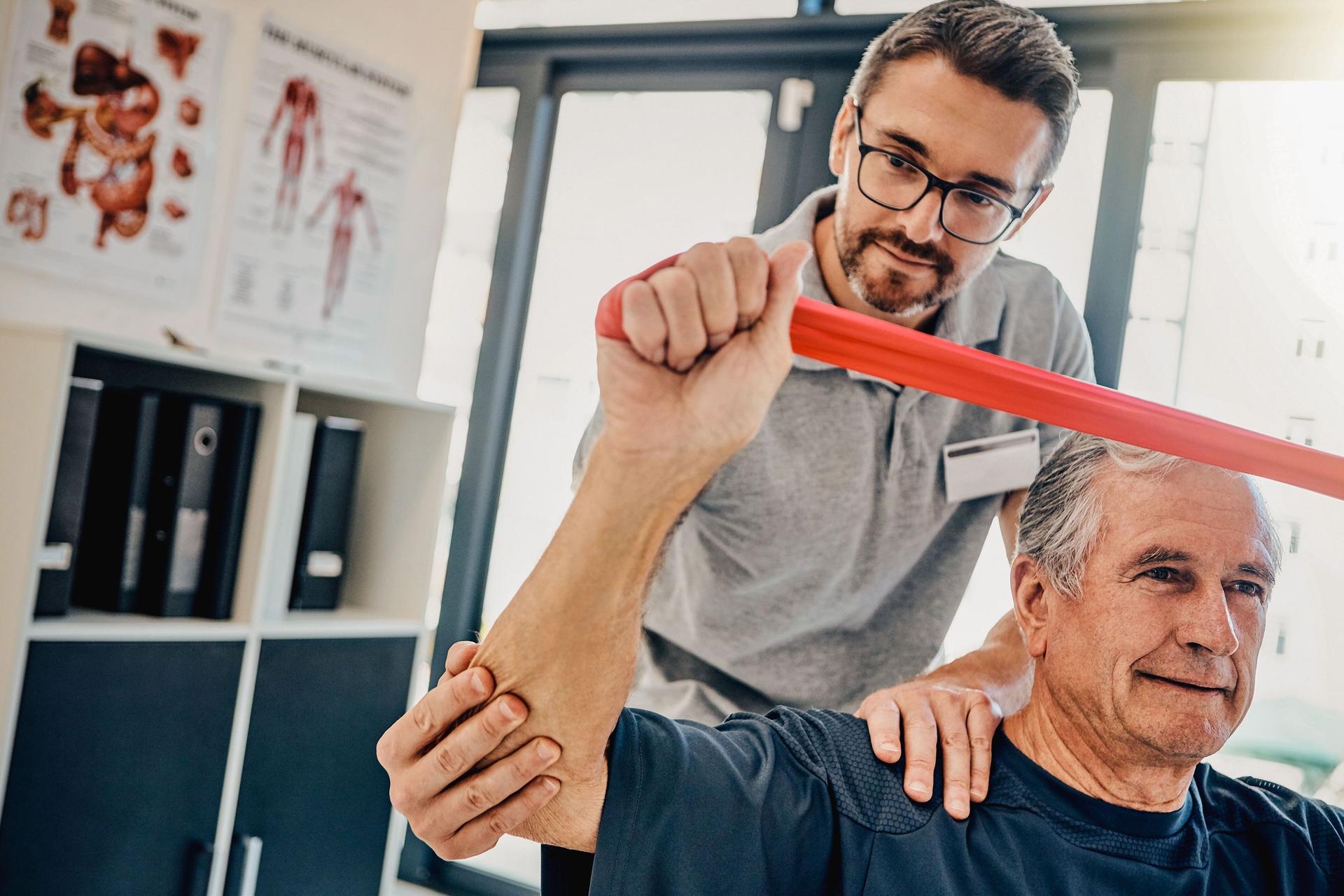 Ein Physiotherapeut hilft einem älteren Patienten, seine Arme mit einen Band zu strecken.