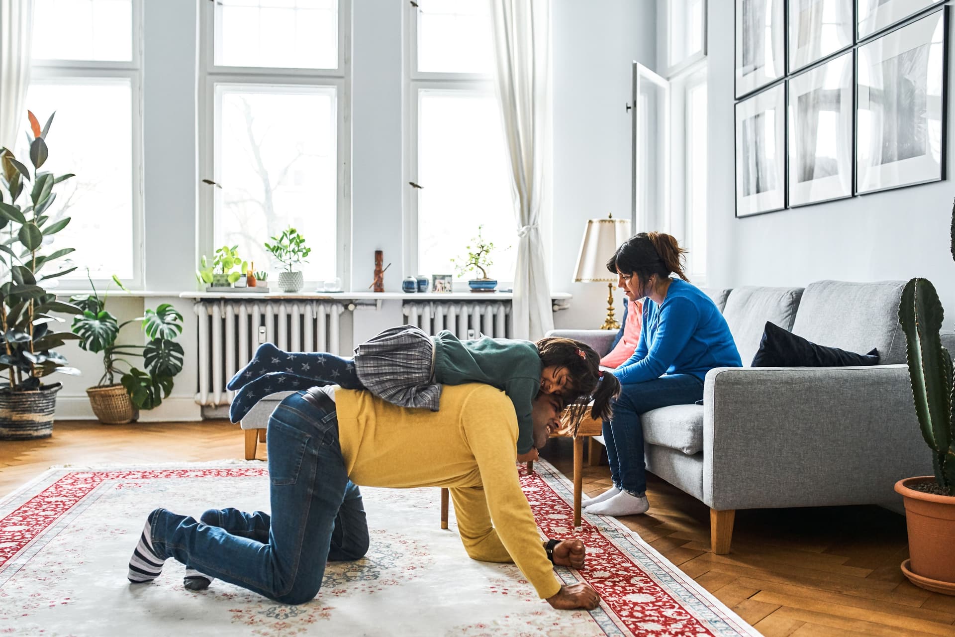 Ein Mann, der auf einem großen Teppich kniet, trägt ein kleines Mädchen auf dem Rücken. Eine Frau sitzt in dem großen hellen Altbauzimmer auf einem Sofa daneben.