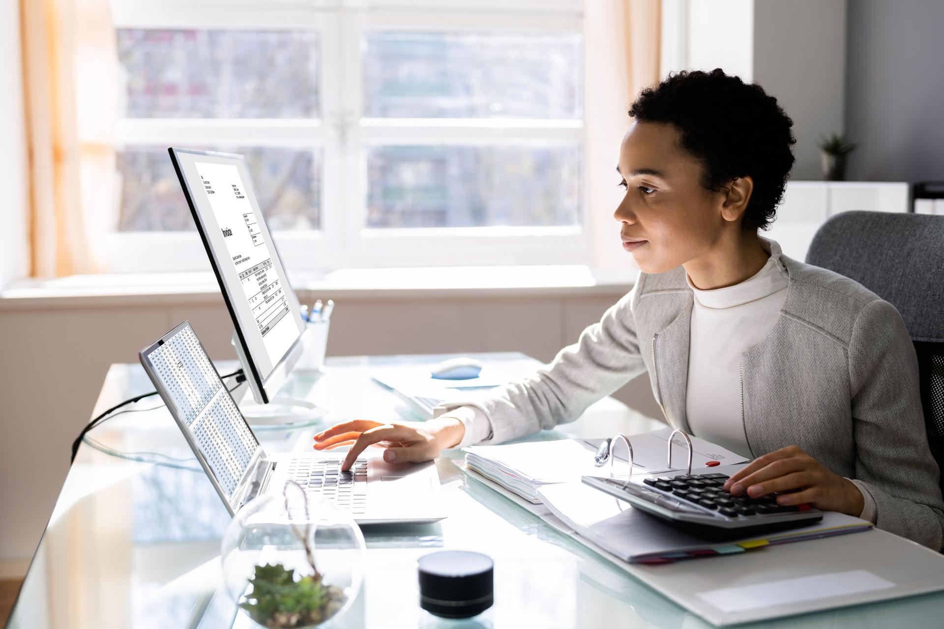 Eine Frau sitzt mit einem Rechner vor einem Laptop und einem Bildschirm. Mit der linken Hand bedient sie den Rechner, mit der rechten Hand tippt sie am Laptop.