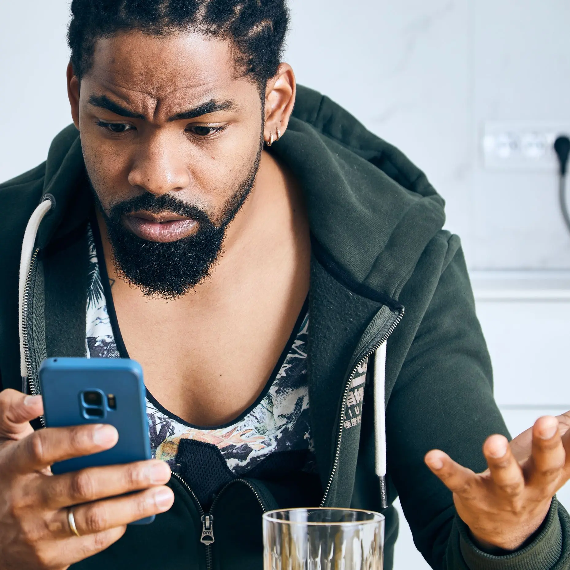 Ein Mann mit Bart sitzt in einer Küche und schaut mit verwirrtem, besorgtem Gesichtsausdruck auf sein Smartphone, während er eine Hand fragend hebt.