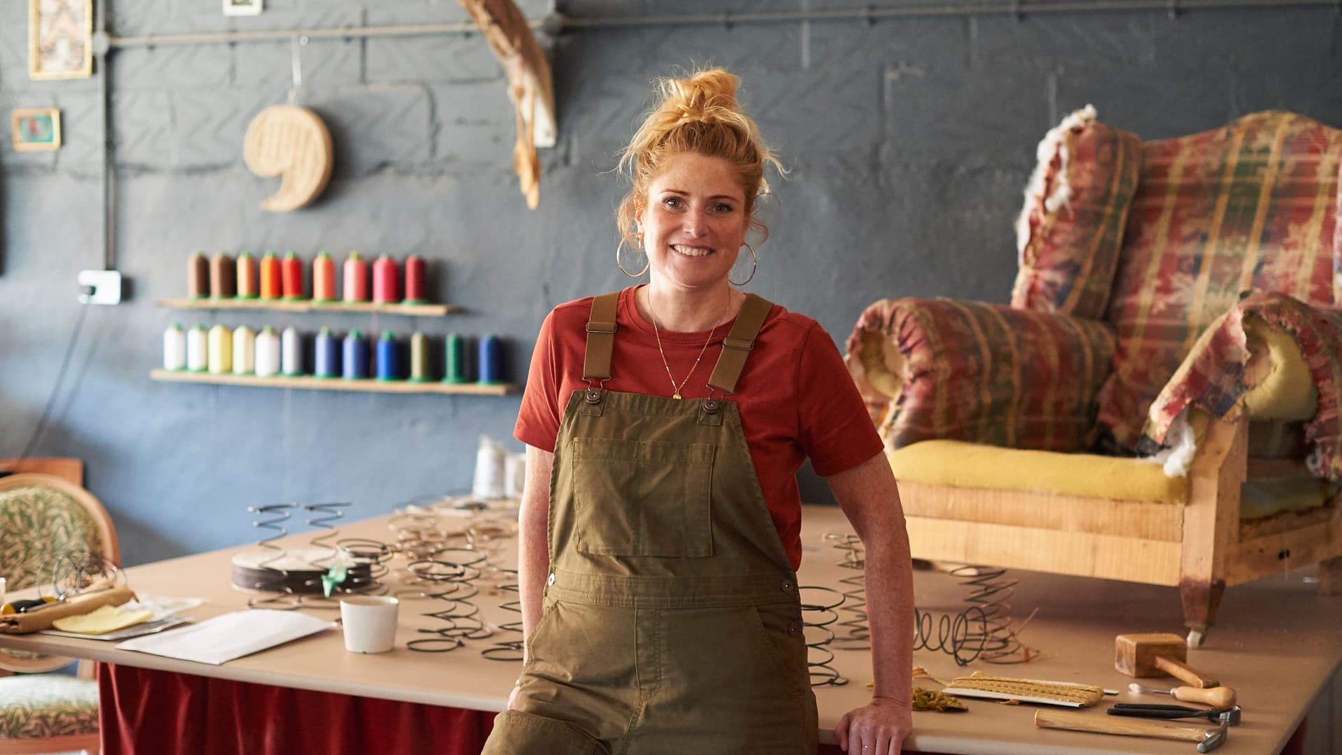 Eine Polsterin steht in ihrer Werkstatt an einen Tisch gelehnt. Im Hintergrund steht ein Sessel zur Reparatur.
