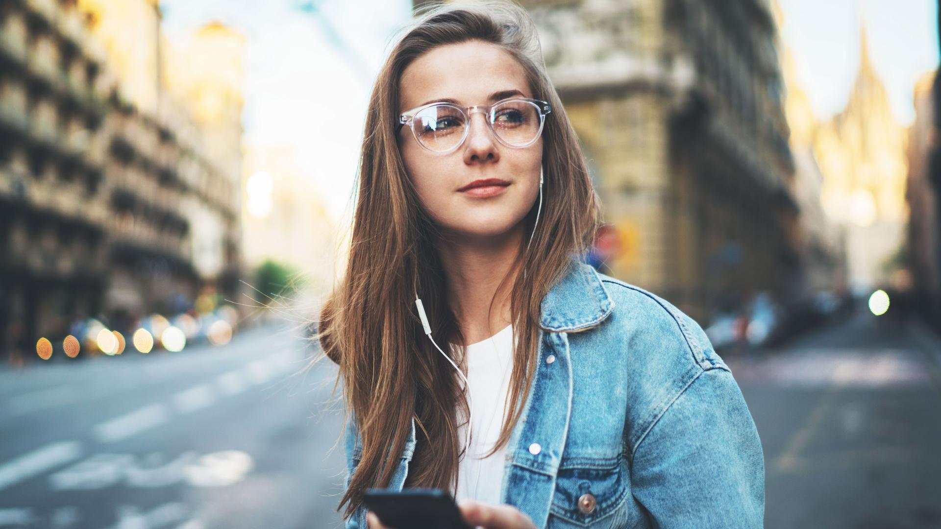 Junge Frau mit Jeansjacke und Brille trägt Kopfhörer und schaut sich im urbanen Raum um.