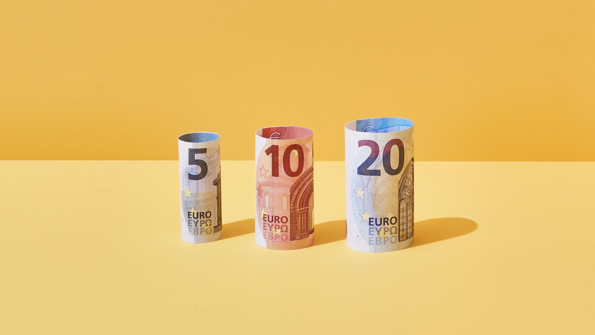 Jeweils ein gerollter 5-Euro, 10-Euro und 20-Euro-Schein stehen nebeneinander vor gelbem Hintergrund