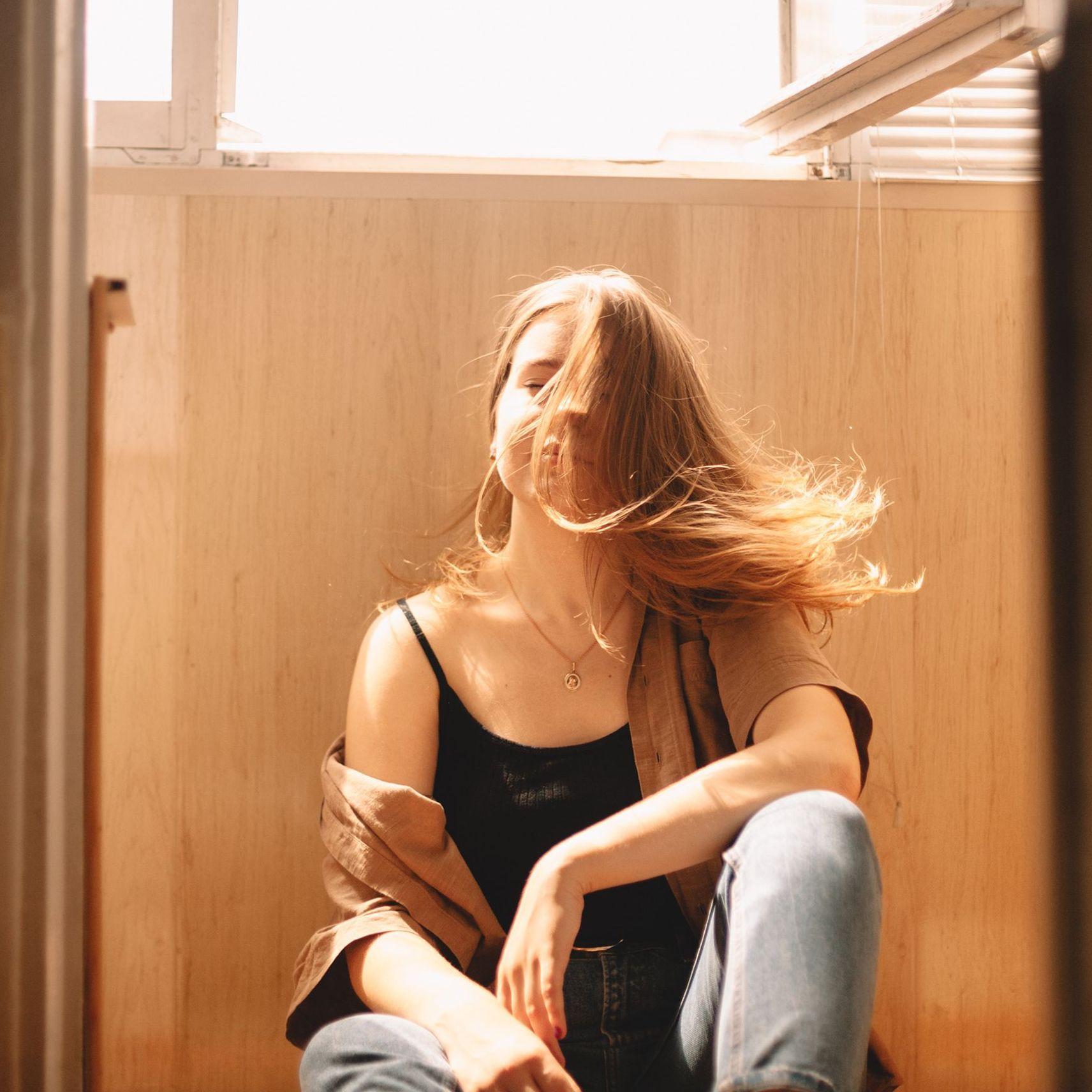 Eine junge, sommerlich gekleidete Frau sitzt zuhause unter einem offenen Fenster und lässt ihre langen Haare im Windzug wehen. 